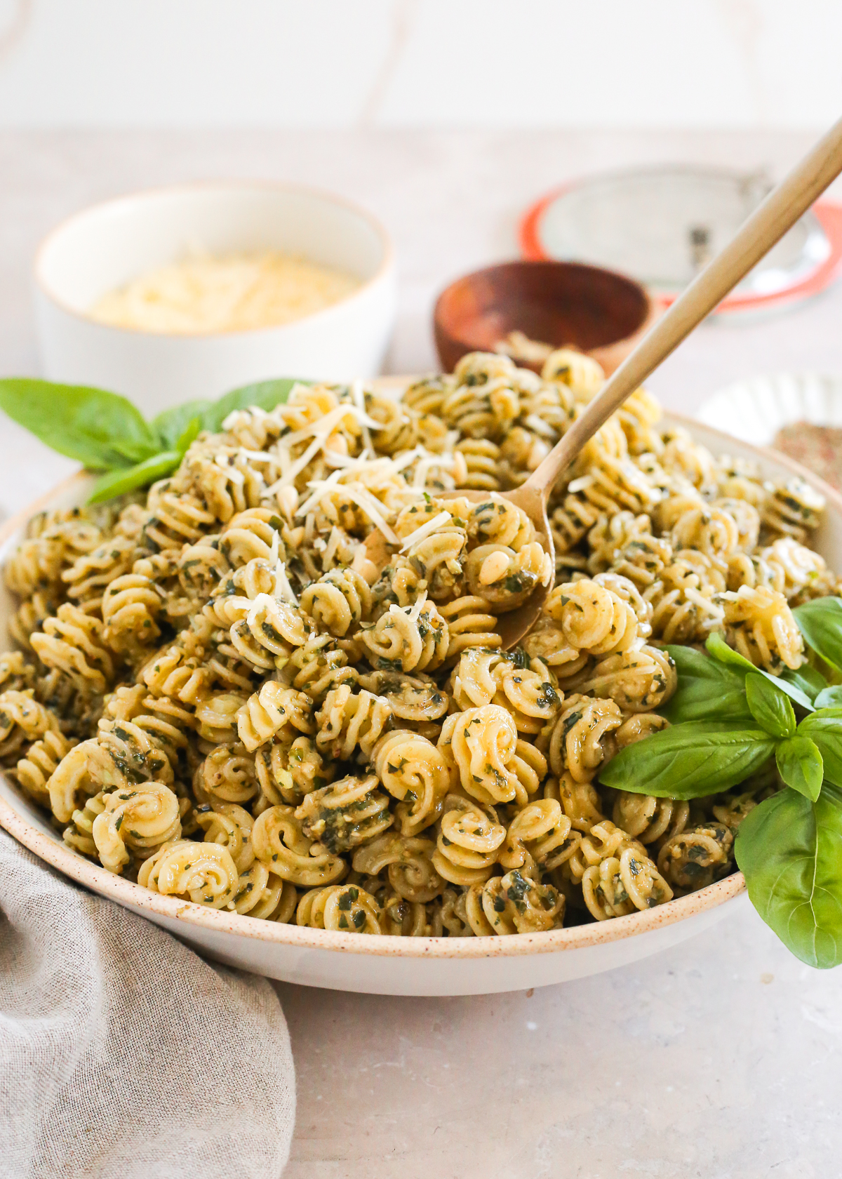 A serving spoon lifts a scoop of basil pesto pasta from a serving bowl on a kitchen countertop.