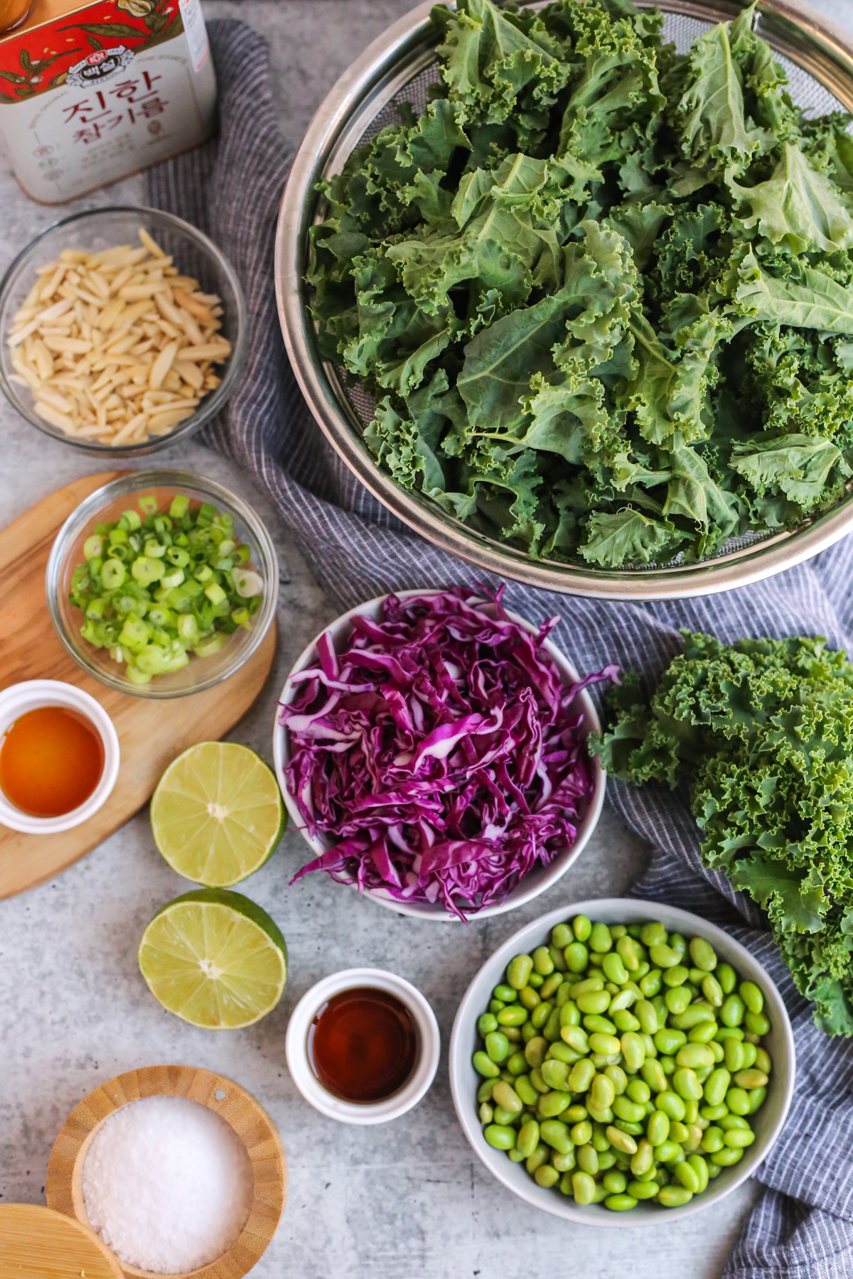 Flatlay view of the ingredients needed to make a massaged kale salad with sesame lime dressing, displayed on a kitchen countertop in various sized prep bowls