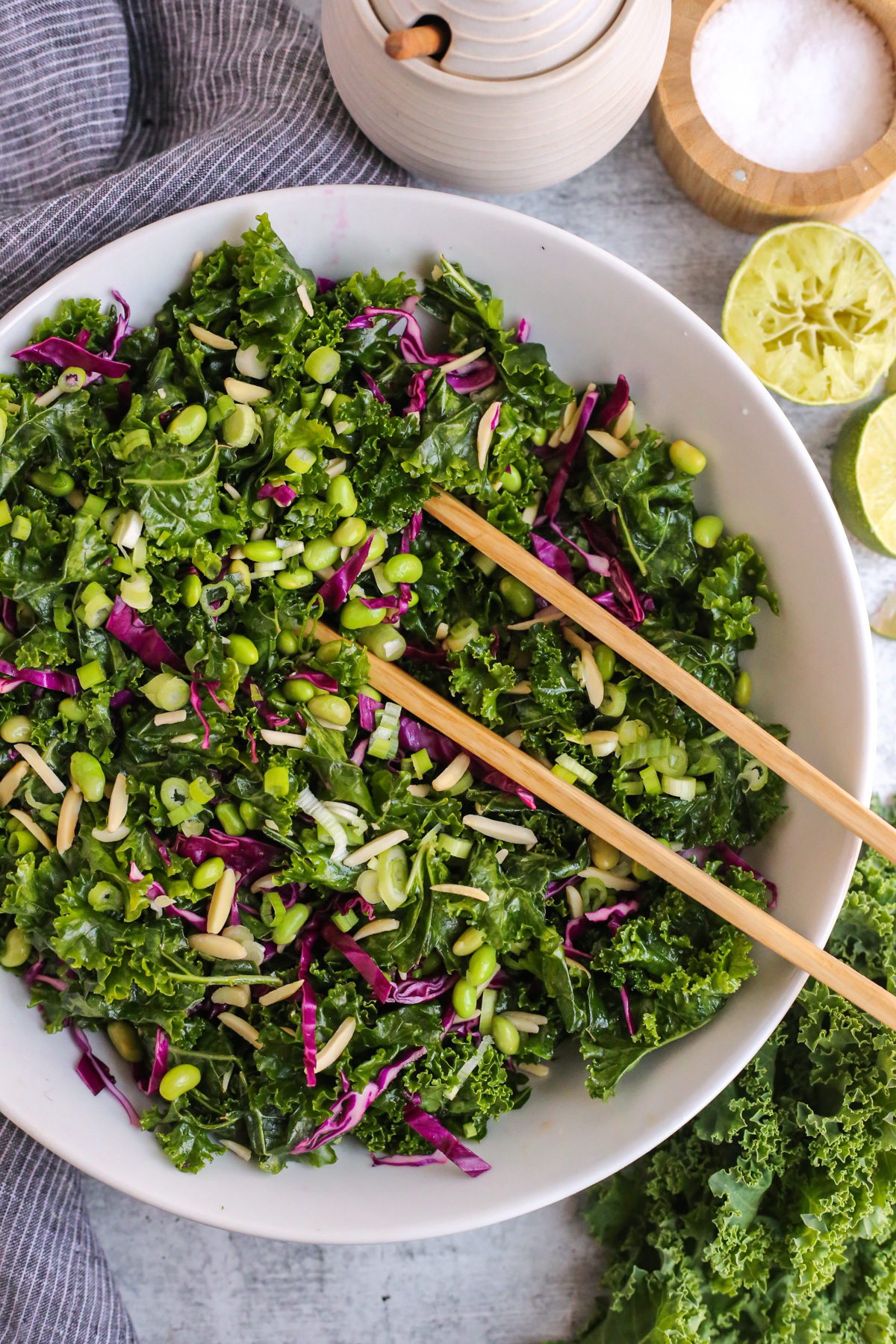 Overhead view of a large white mixing bowl filled with a massaged kale salad, shelled edamame, shredded red cabbage, slivered almonds, sliced green onions, and a light sesame-lime dressing, with a pair of wooden salad tongs ready to serve it
