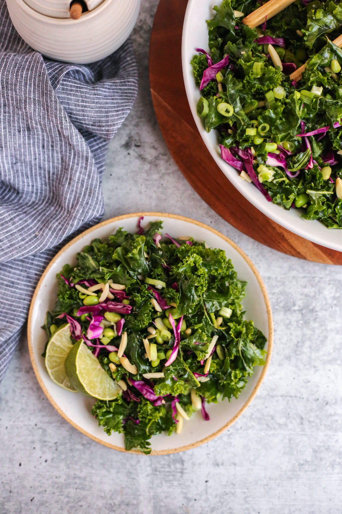 Overhead view on a kitchen countertop, showing a side salad served from a larger serving bowl. The salad is colorful with raw, massaged kale, lime wedges, red cabbage, edamame, slivered almonds, and sliced green onions. It's lightly dressed with a sesame-lime dressing and it looks fresh and appetizing
