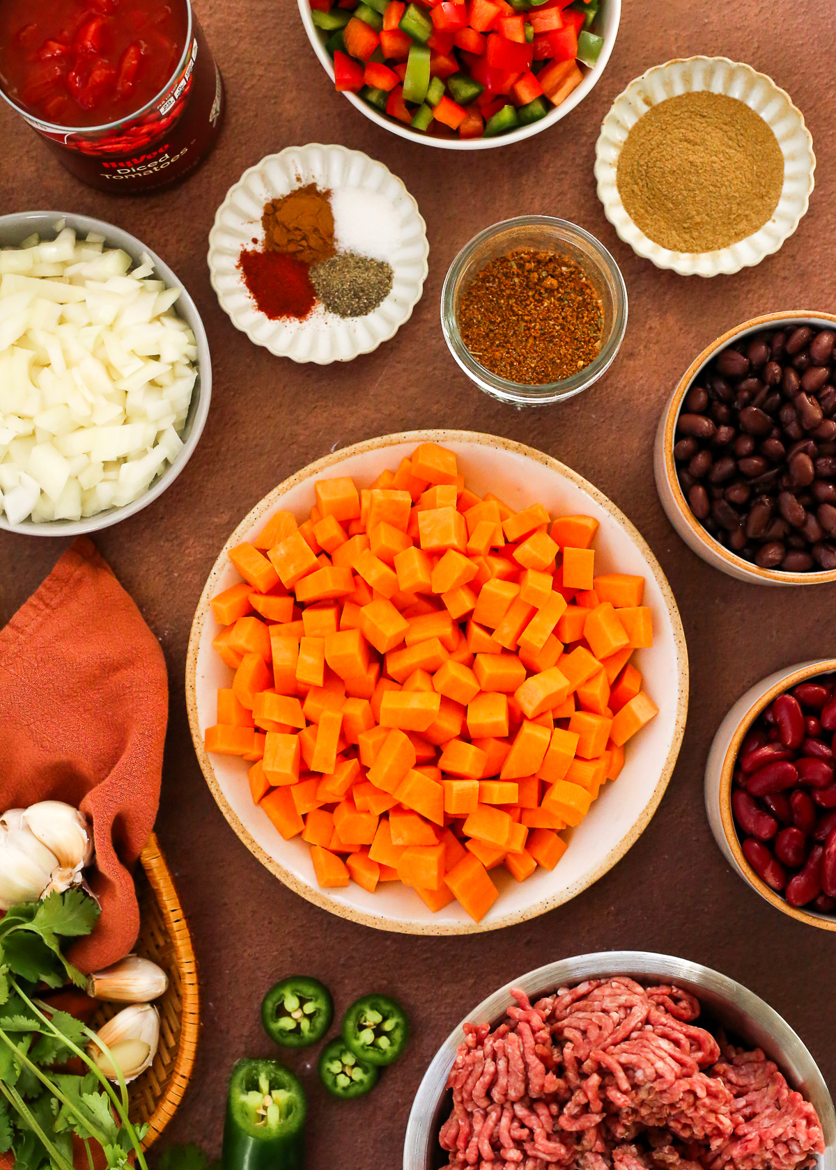 An assortment of ingredients to make a sweet potato chili recipe is displayed on a dark stone countertop.
