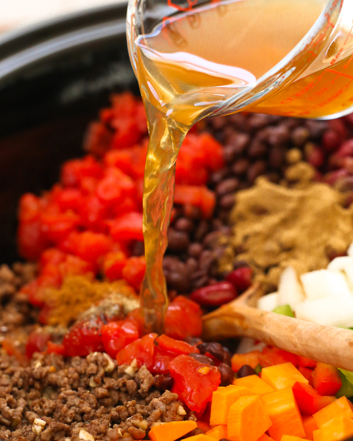 A clear glass measuring cup pours vegetable broth into the bowl of a slow cooker.