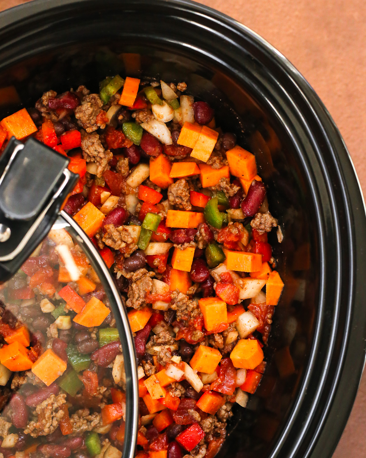 A mixutre of sweet potato chili in a slow cooker with the lid positioned as if about to be sealed and simmered. 