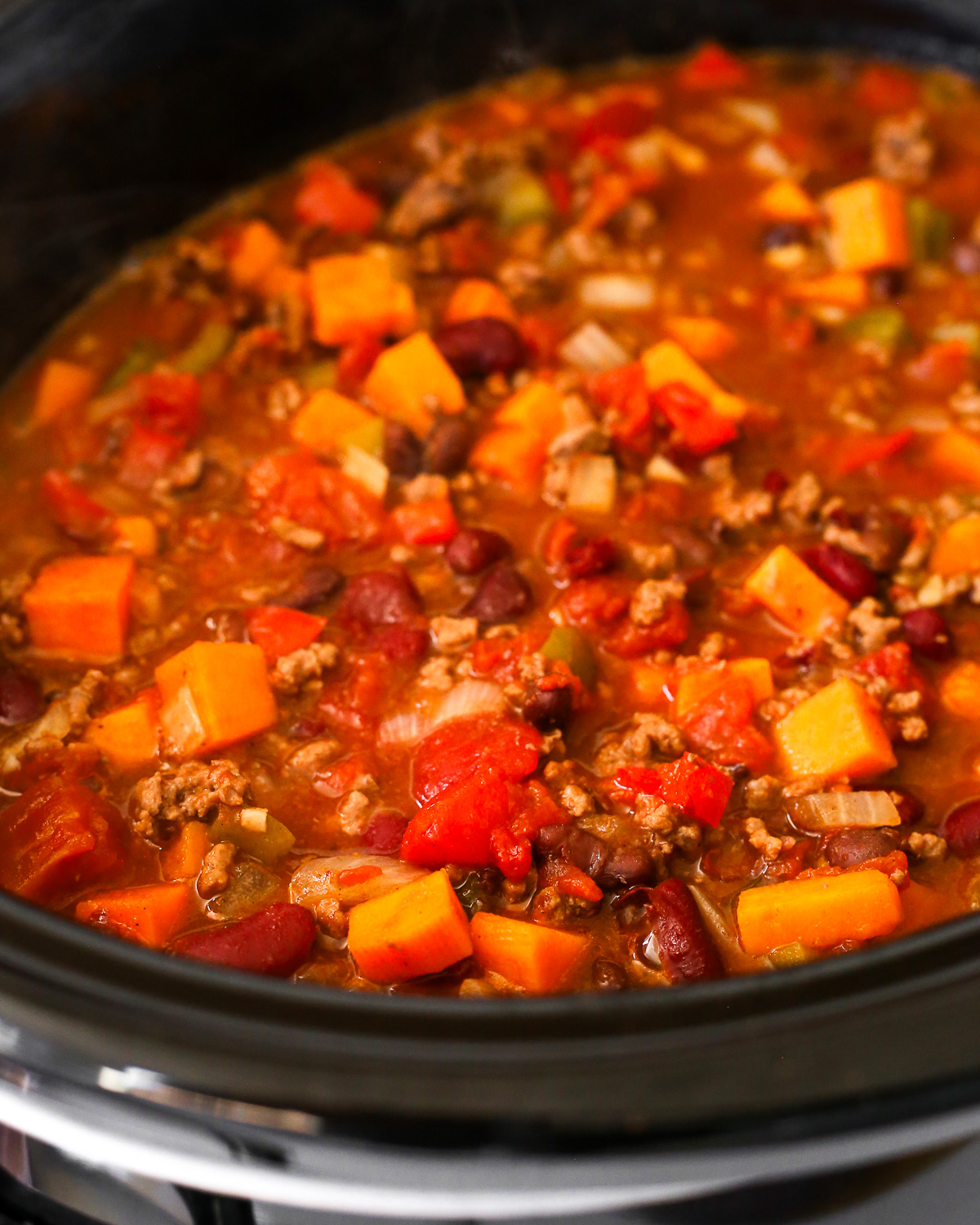A close view of cooked chili in a slow cooker, showing a variety of beans, ground beef, peppers, onions, and sweet potatoes. 