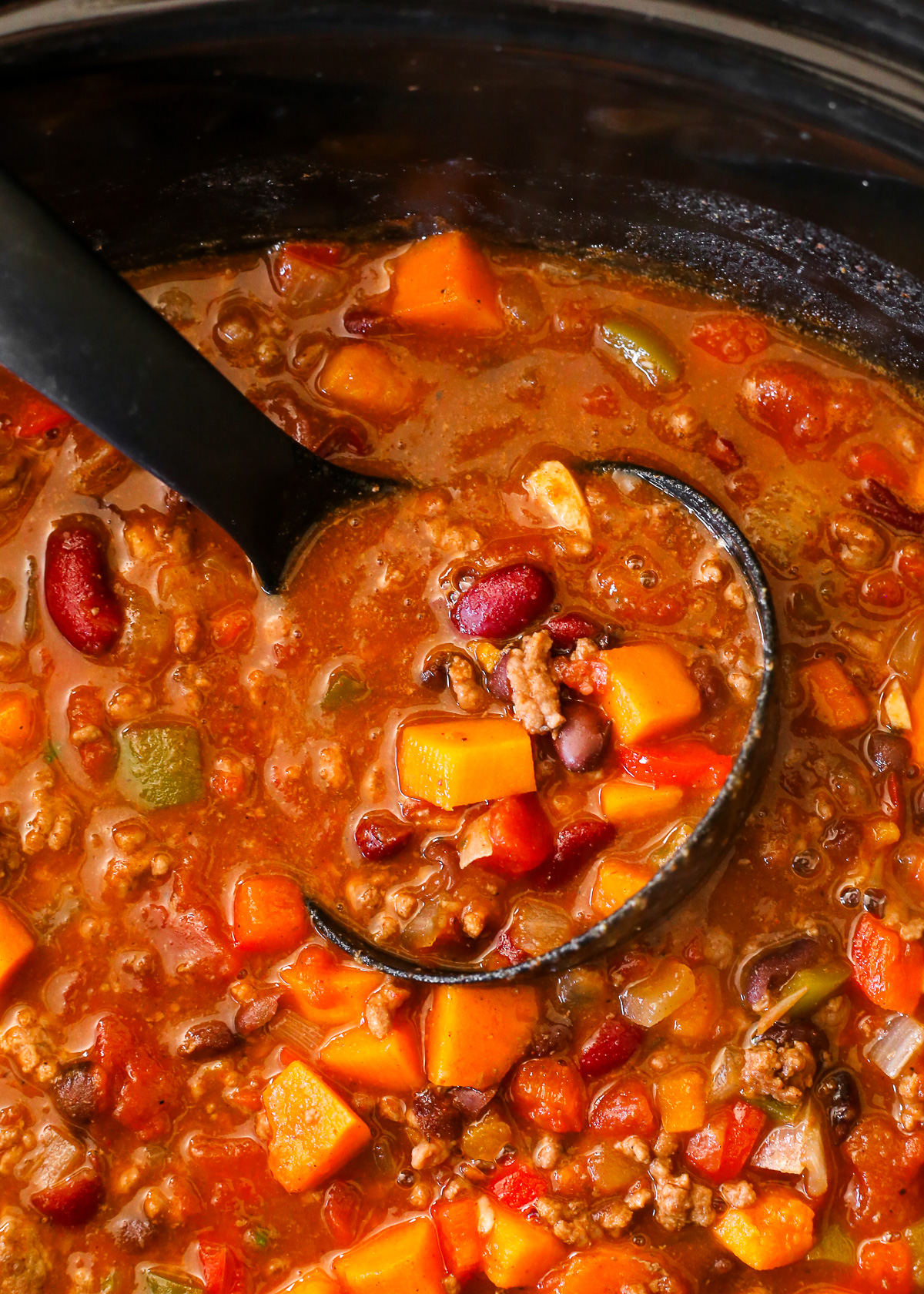 A black ladle scoops a spoonful of chili from a slow cooker, showing a variety of tomatoes, ground beef, beans, peppers, onions, and sweet potatoes.