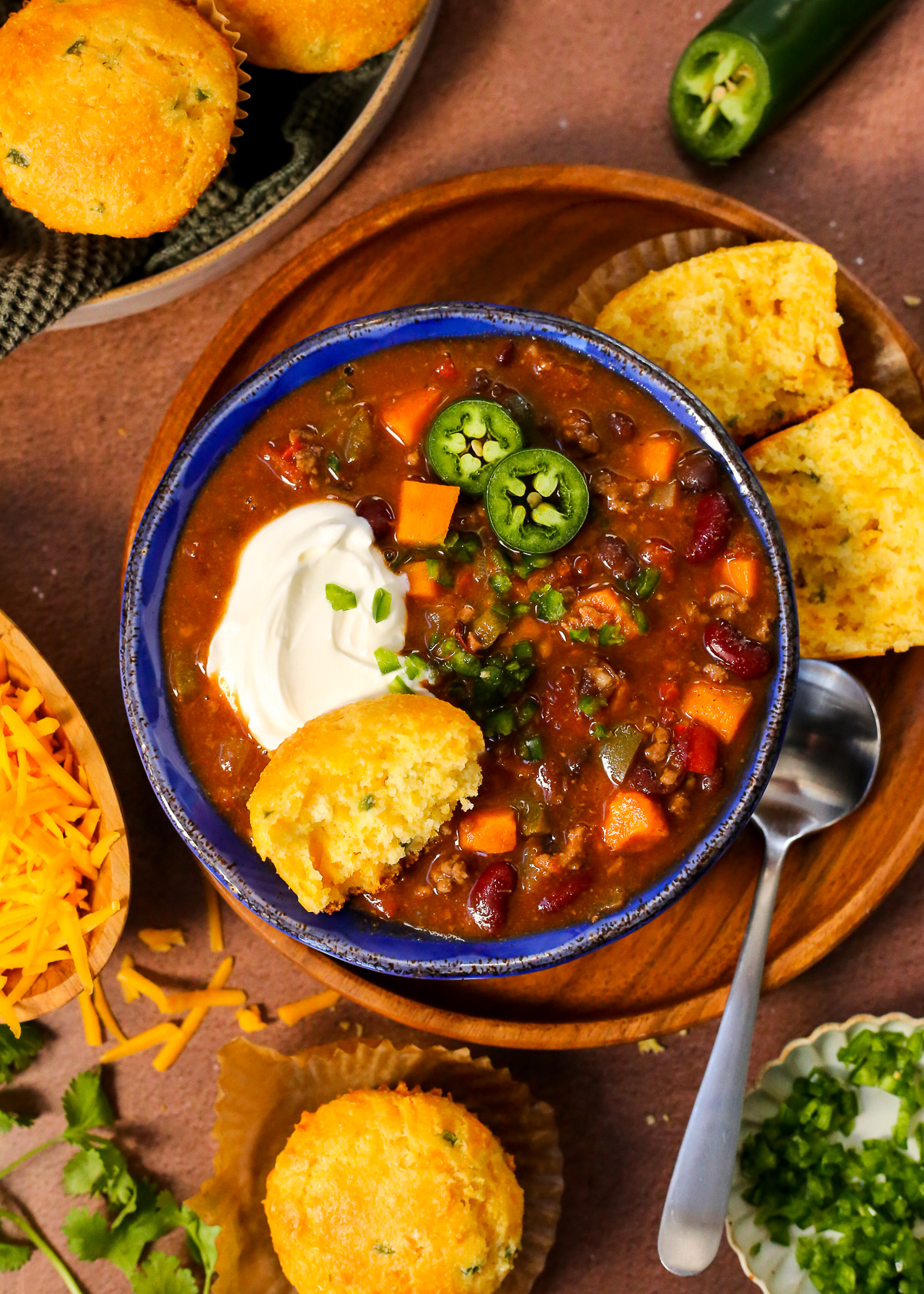 A blue ceramic bowl filled with a sweet potato chili, topped with a dollop of sour cream, minced jalapenos, and a cornbread muffin. 