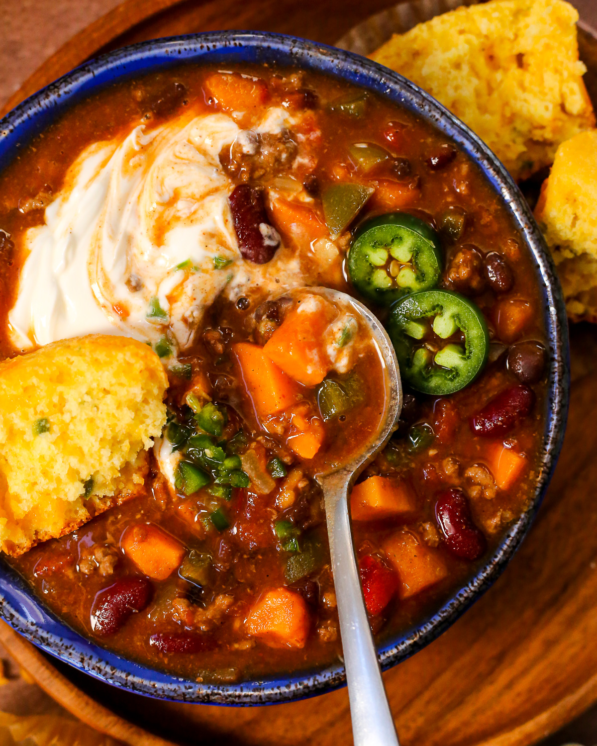 A bowl of sweet potato chili topped with sour cream, minced jalapenos, half of a cornbread muffin, and a spoon gently swirling the sour cream into the broth.