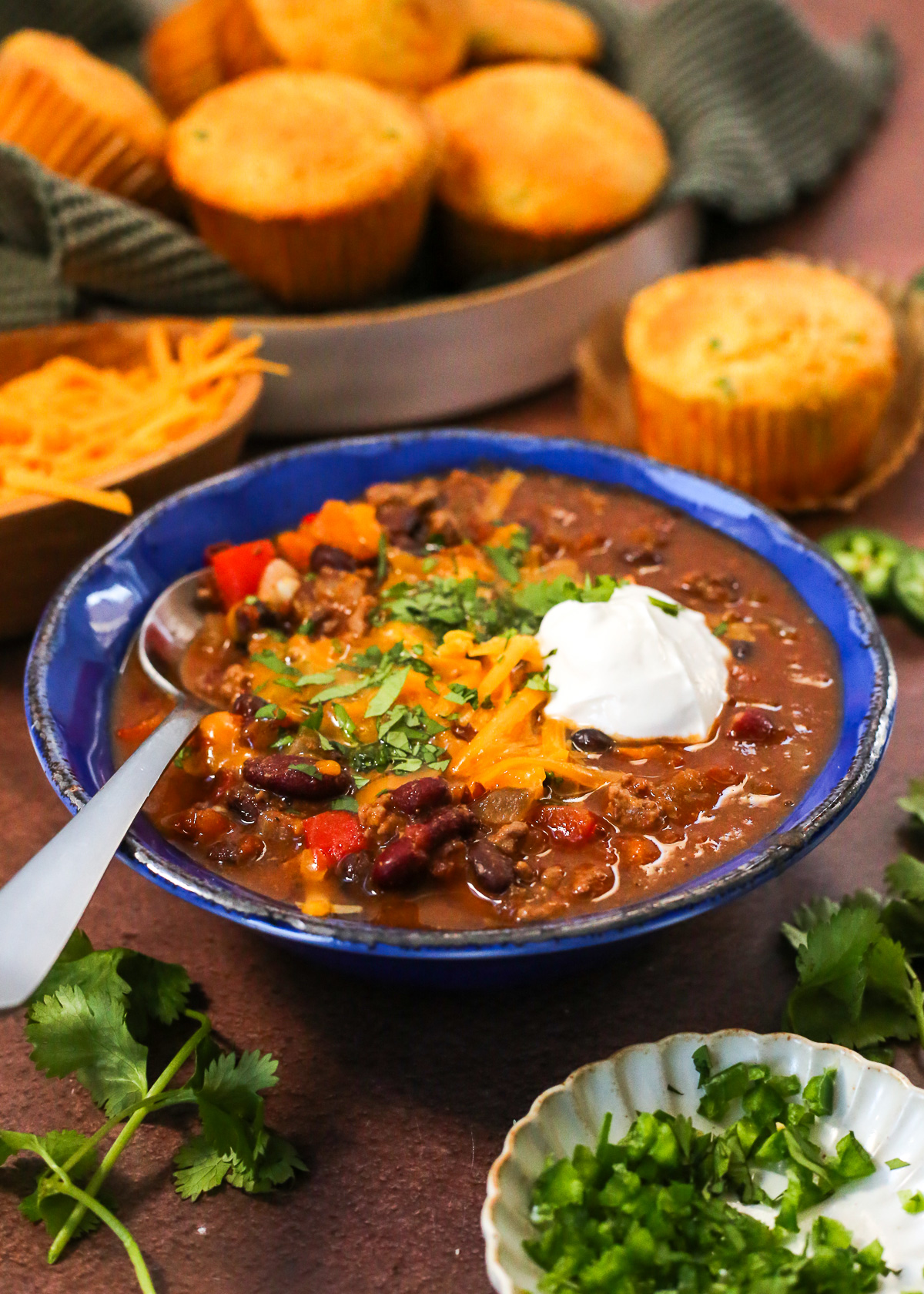 A blue ceramic bowl of sweet potato chili displayed on a kitchen countertop, topped with melted cheddar cheese, cilantro, and a scoop of sour cream. 
