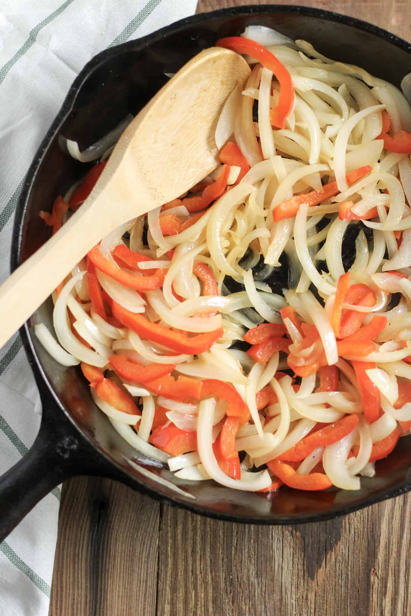 Overhead view of sliced peppers and onions in a cast iron skillet, stirred with a wooden spoon