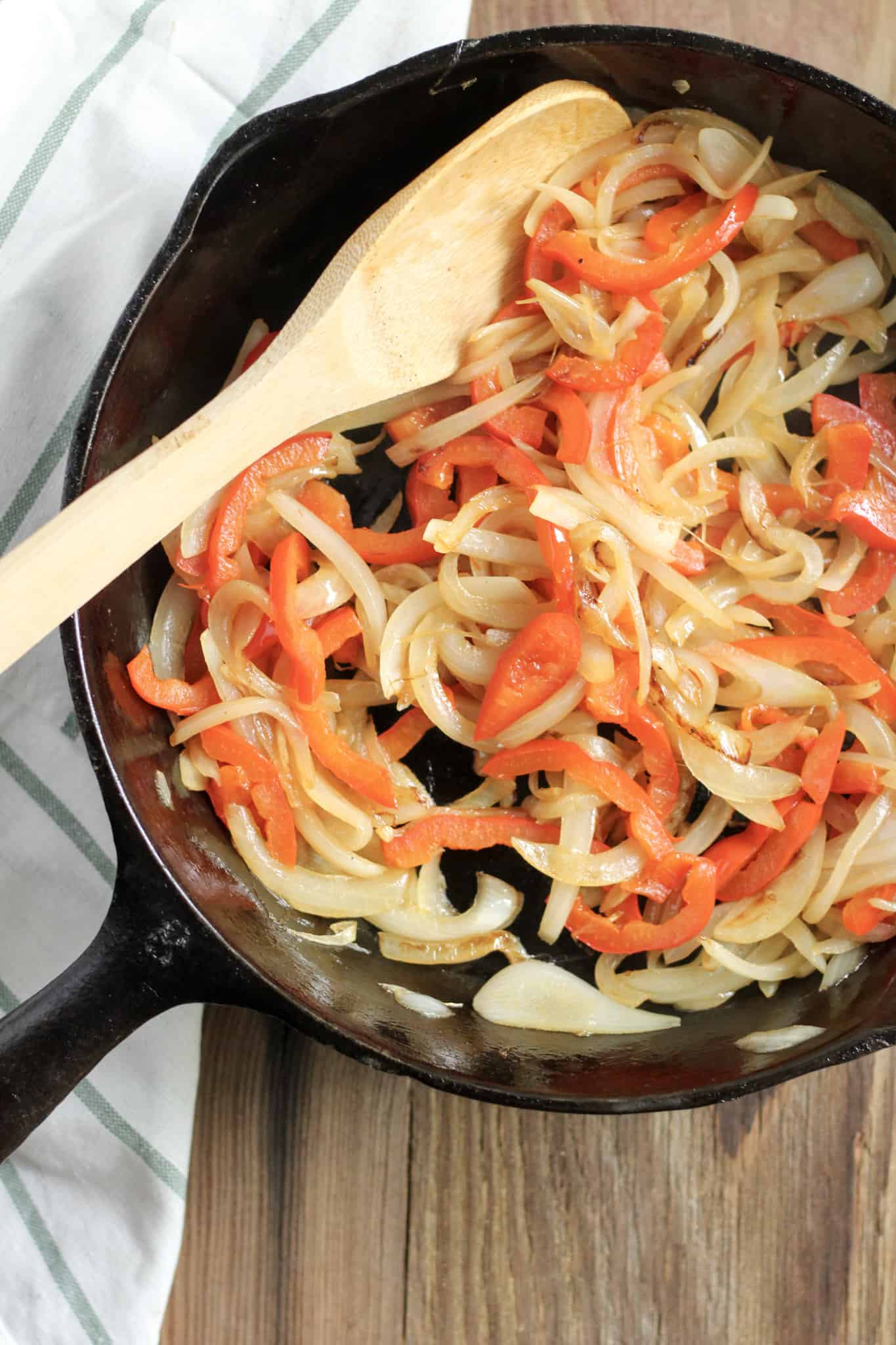 Overhead view of a cast iron skillet with cooked peppers and onions, with a wooden spoon resting on the side of the skillet