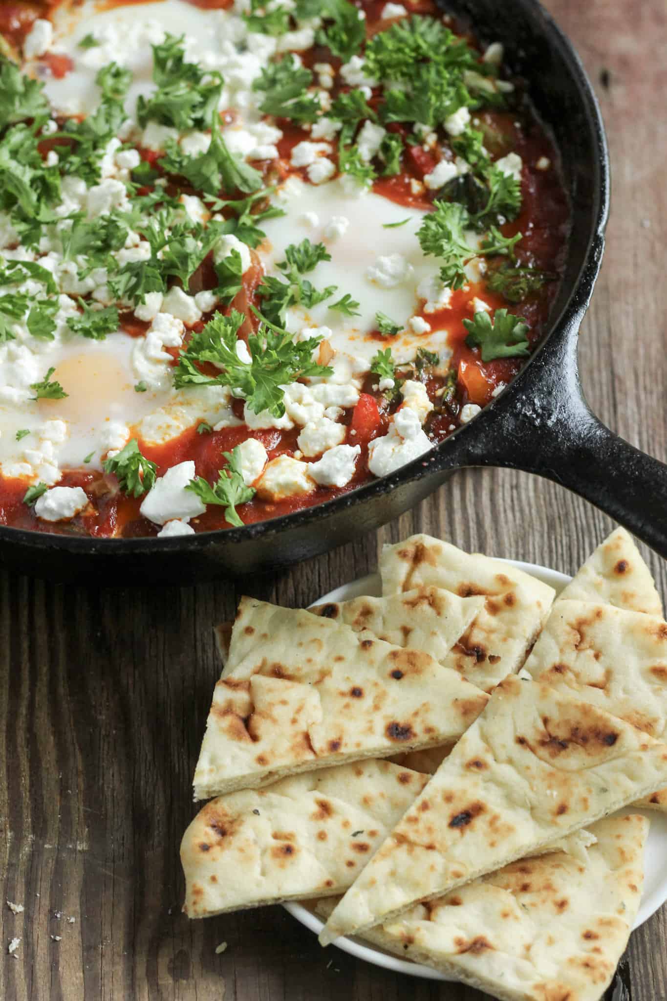 Cast iron skillet with an easy shakshuka recipe on a wooden table with a serving plate of sliced naan