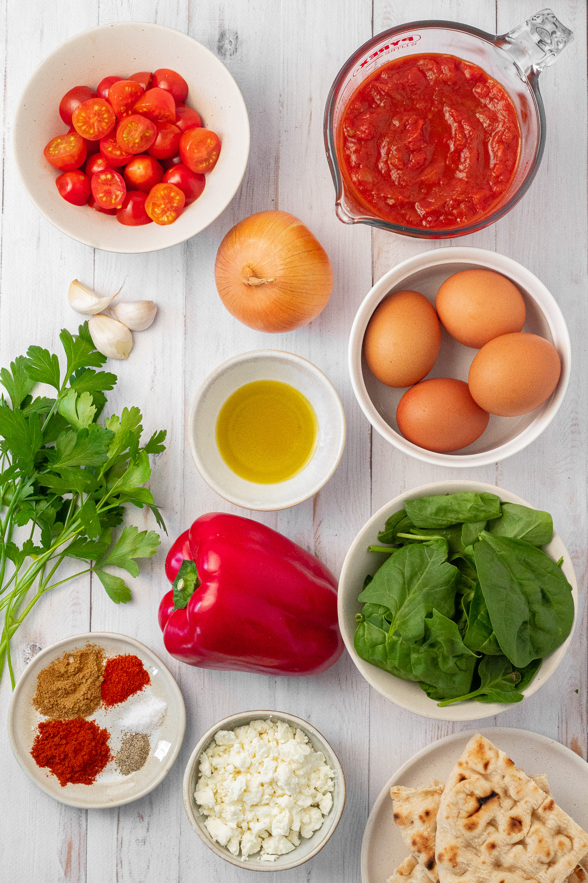 An assortment of ingredients needed to make a beginner-friendly shakshuka recipe, including fresh spinach and parsley, marinara sauce, cherry tomatoes, garlic and onion, red bell pepper, feta cheese, pita bread, and spices. 