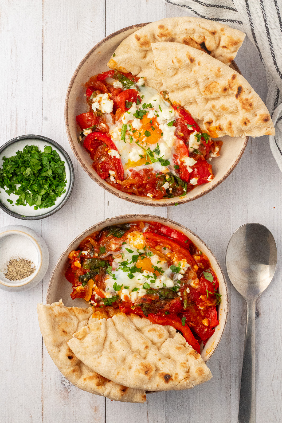 Two bowls of shakshuka served with sliced pita bread, topped with chopped parsley and feta cheese crumbles.