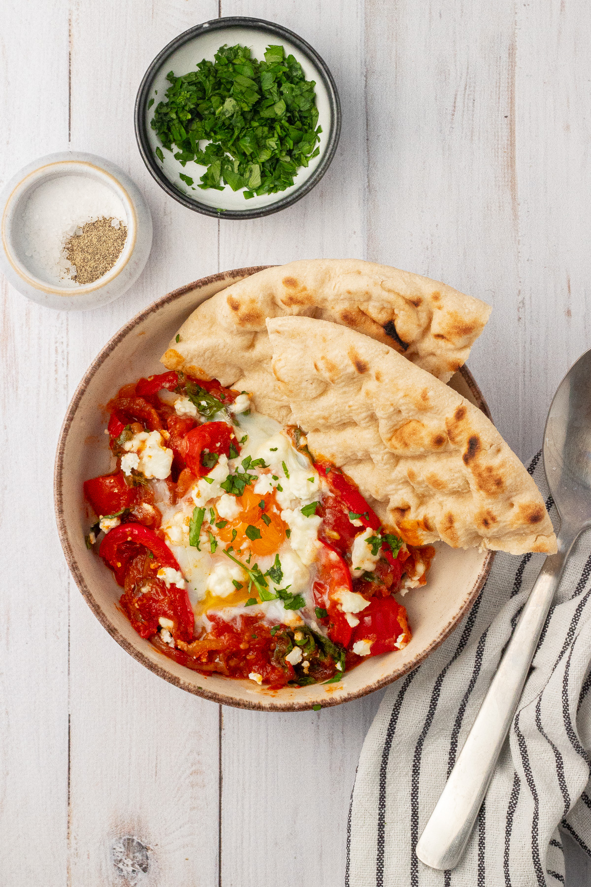 A bowl of shakshuka made with marinara sauce is served with two slices of pita bread, chopped parlsey, and extra salt and pepper on the side. 