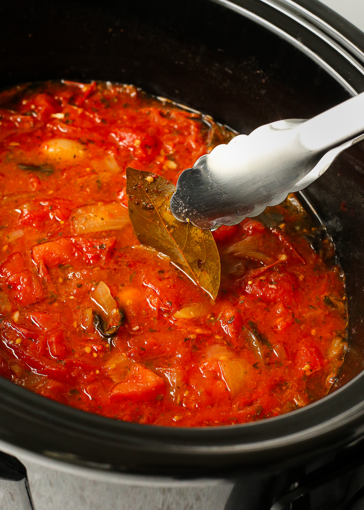 A pair of metal tongs lifts a used bay leaf from the marinara sauce that's been cooking for hours in a slow cooker.