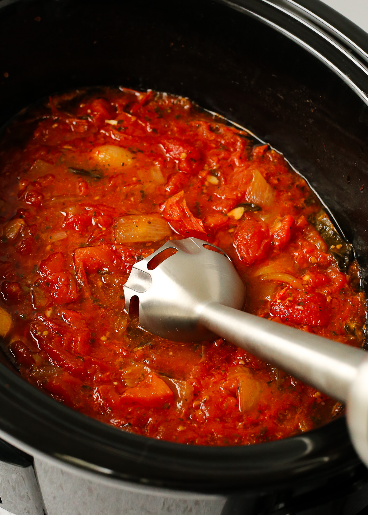 An immersion blender rests on the side of a slow cooker, prepared to blend the marinara sauce that's been stewing.