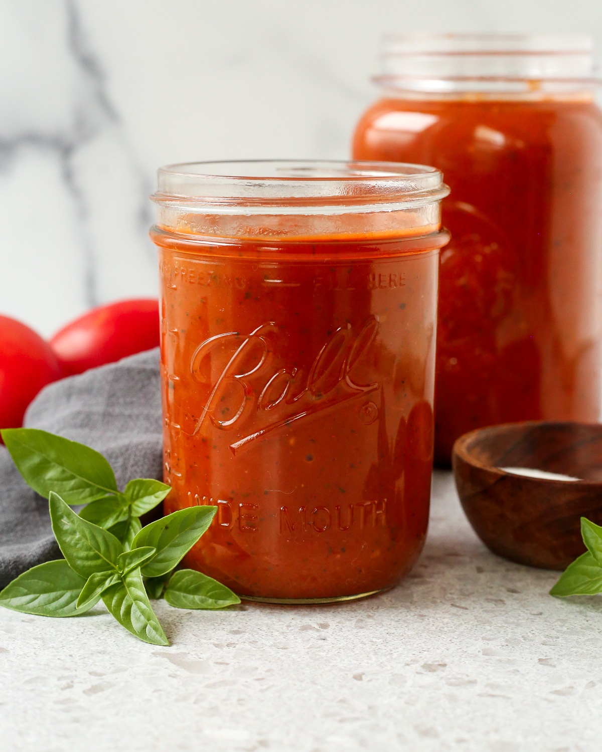 Two large storage jars full of slow cooker marinara sauce displayed on a kitched countertop surrounded by fresh basil leaves.