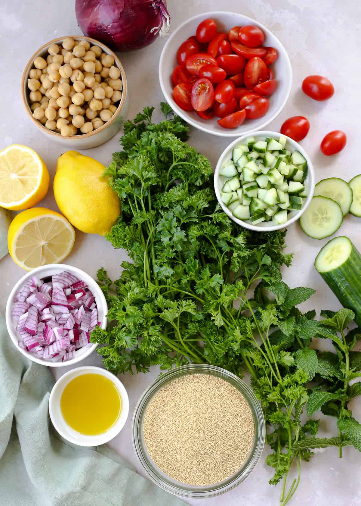 An assortment of fresh ingredients displayed on a kitchen countertop, including curly parsley, fresh mint, cherry tomatoes, canned chickpeas, a lemon, diced red onion, diced cucumber, and uncooked amaranth.