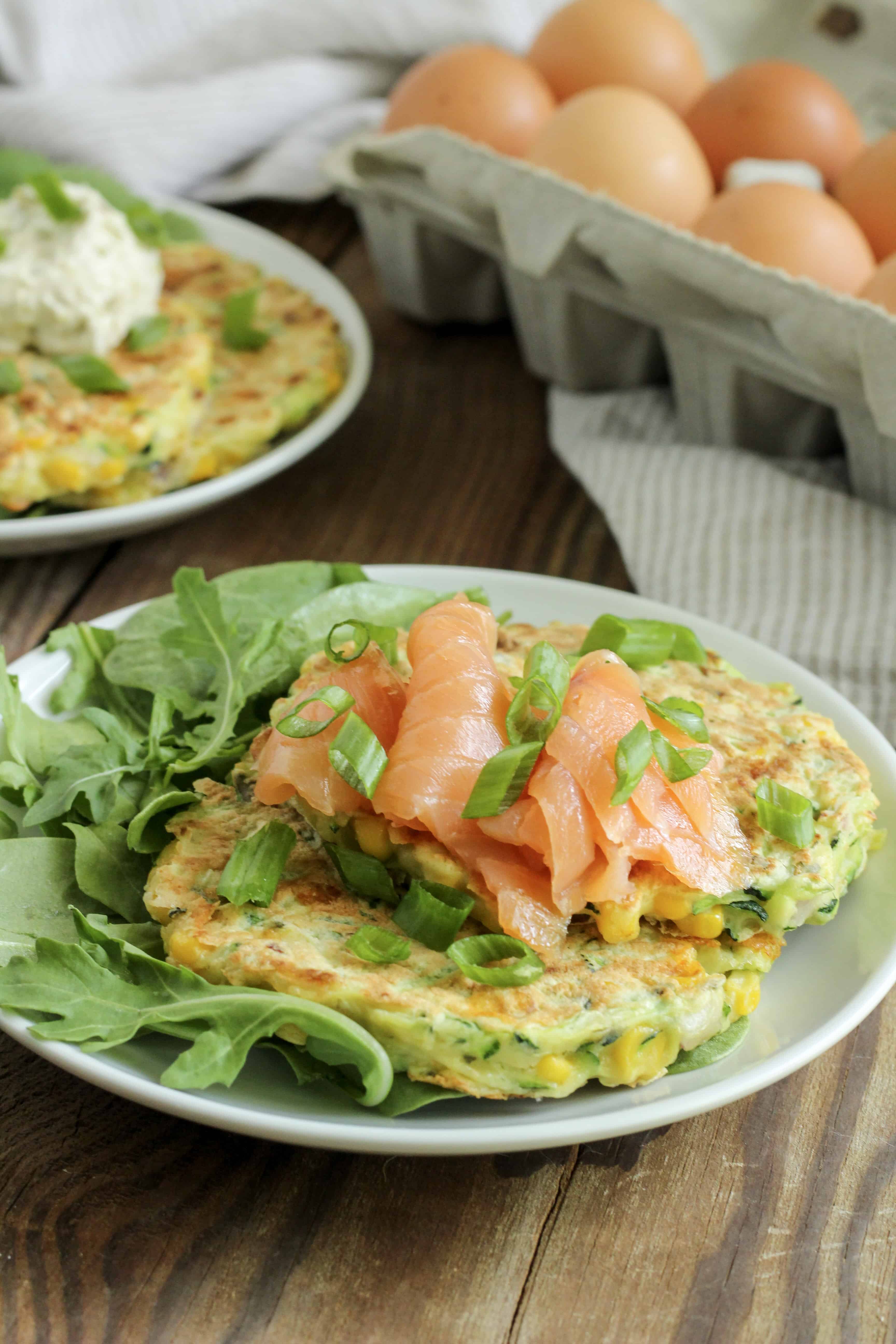 Image of two savory zucchini pancakes served on a small white plate, with a side of salad greens and topped with several slices of smoked salmon