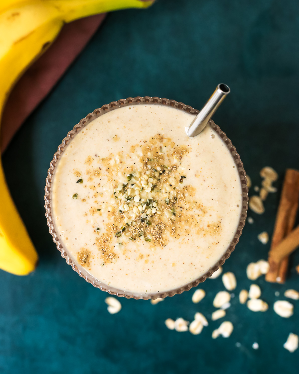 Overhead view of a banana chai smoothie in a clear glass, topped with a dusting of chai spice and a short metal straw resting on one side of the glass. 
