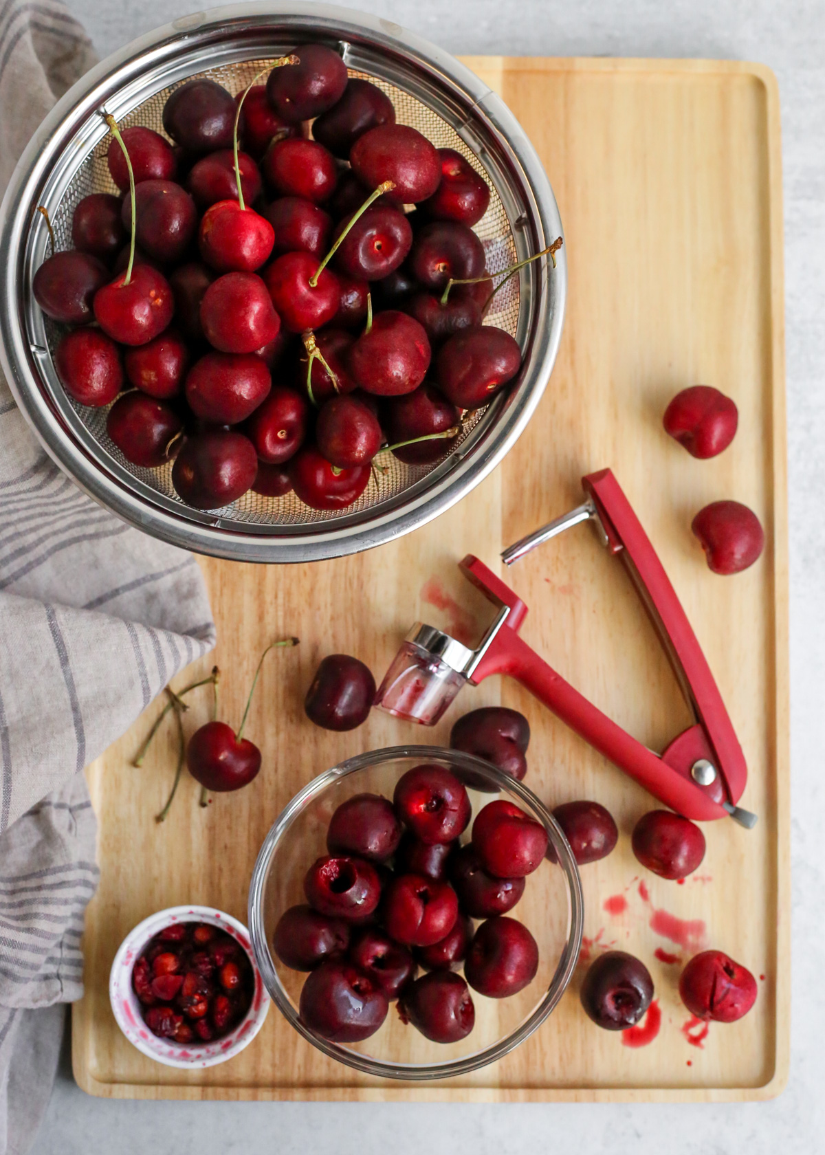 A large colander full of sweet cherries is displayed on a wooden cutting board with a cherry pitter and a bowl of cherries that have been freshly pitted