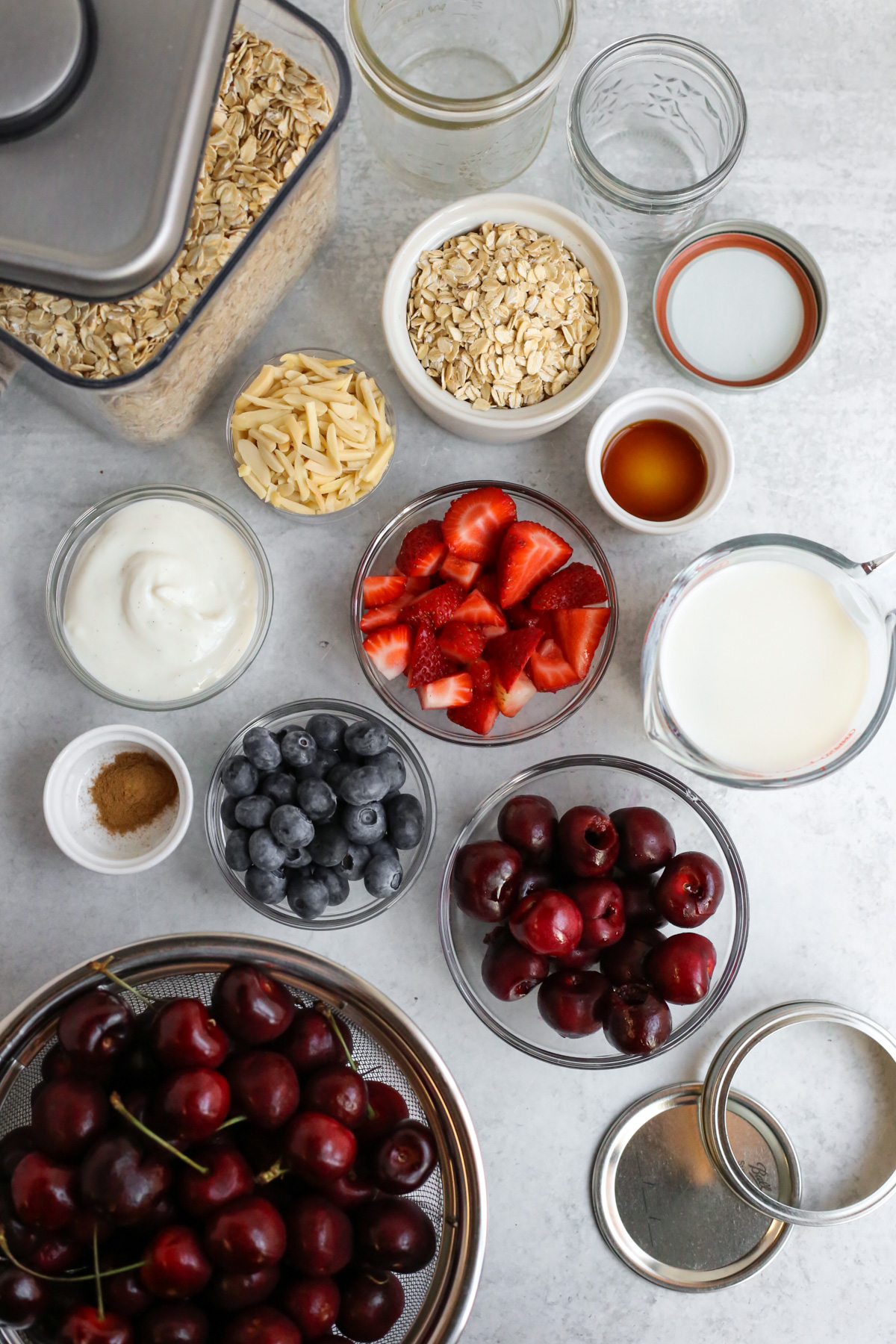 Overhead view of the ingredients needed to make overnight oats, arranged in small jars and including fresh strawberries, blueberries, and cherries, along with a measuring cup on milk, yogurt, vanilla extract, cinnamon, and rolled oats