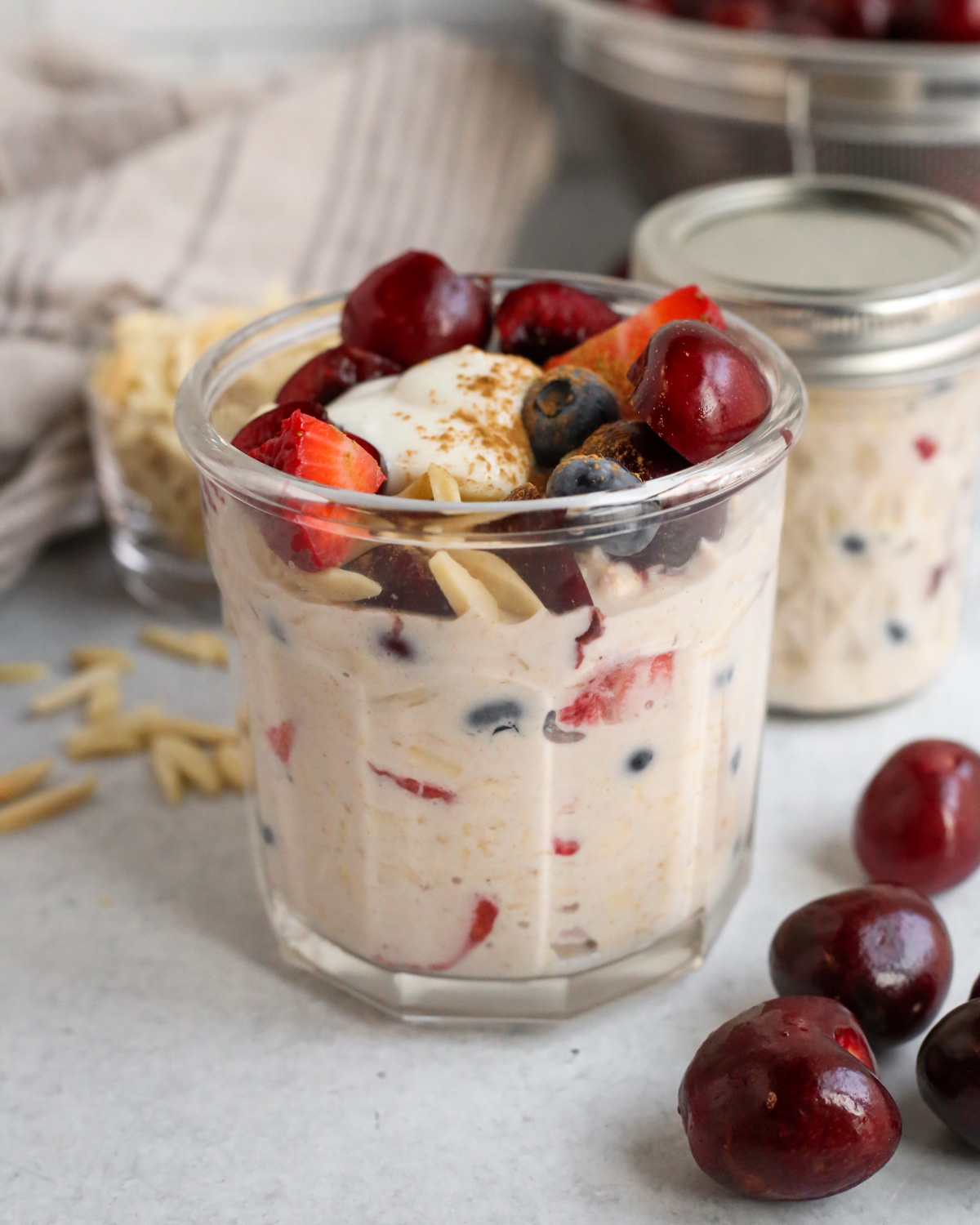 A clear glass jar on a kitchen countertop filled to the top with overnight oats, topped with fresh cherries, strawberries, blueberries, slivered almonds, a dollop of vanilla yogurt, and cinnamon