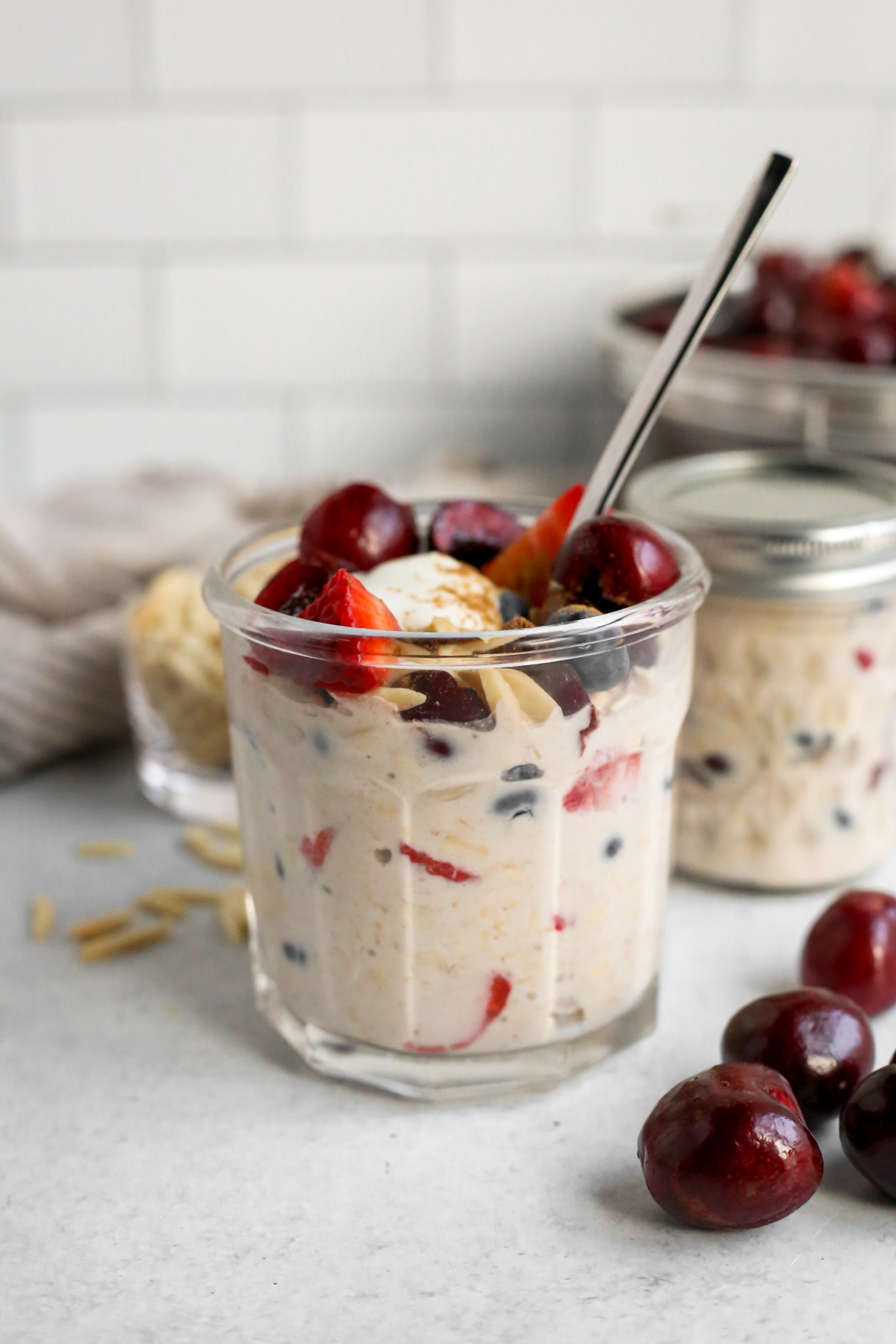 Styled jar of Cherry Overnight Oats, topped with fresh berries and almonds, displayed on a kitchen countertop with fresh cherries in the background and a silver breakfast spoon 