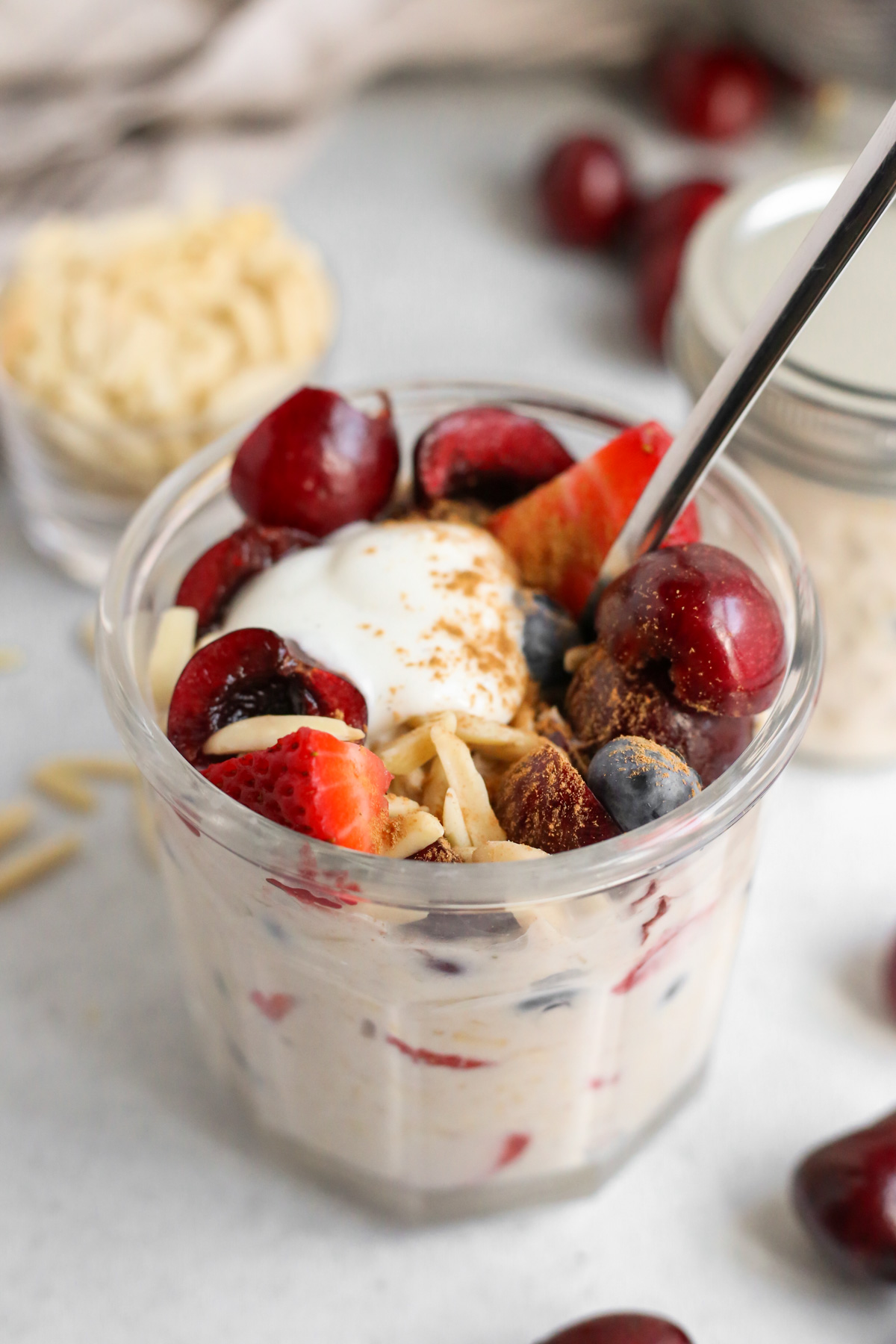 Close up view of Berry Cherry Almond Overnight Oats in a serving jar, topped with extra fruit, almonds, yogurt, and cinnamon with a spoon placed in the jar as if ready to take a bite