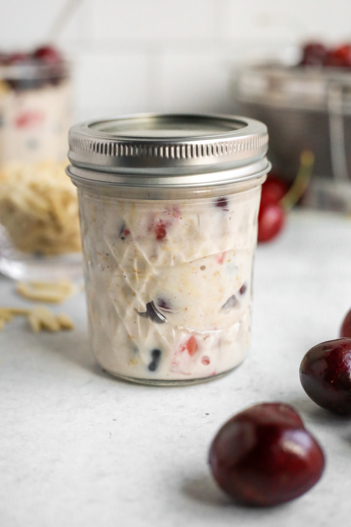 A small mason jar with the lid fastened sits on a kitchen countertop. It's filled with Berry Cherry Almond Overnight Oats, with pieces of the colorful fruit visible in the mixture