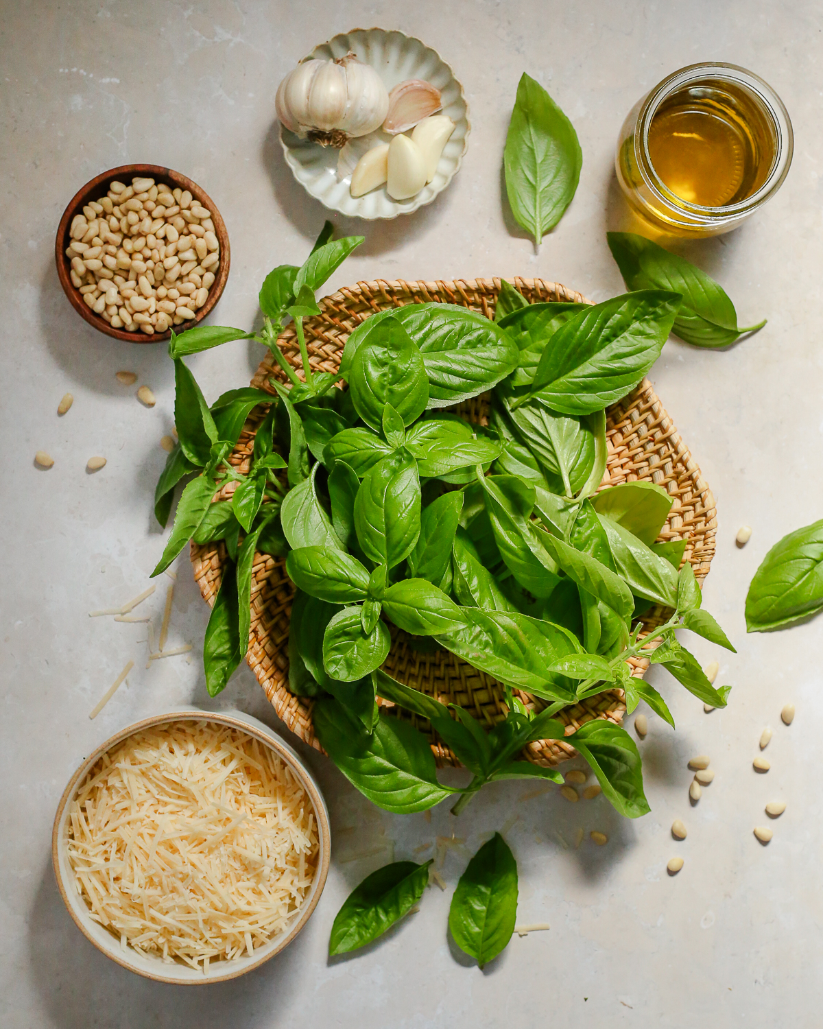 An assortment of ingredients needed to make a basil pesto sauce, including fresh basil leaves, shredded parmesan cheese, pine nuts, garlic, and olive oil.