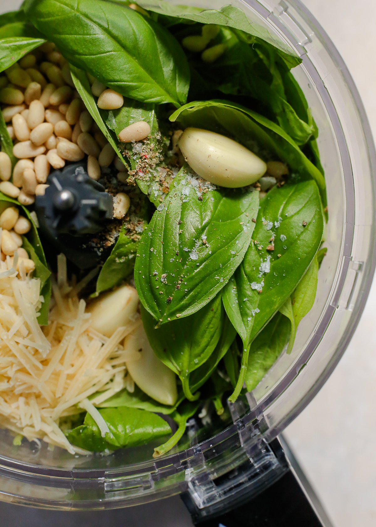 A view into the bowl of a food processor containing fresh basil, garlic cloves, parmesan cheese, pine nuts, and salt and pepper.
