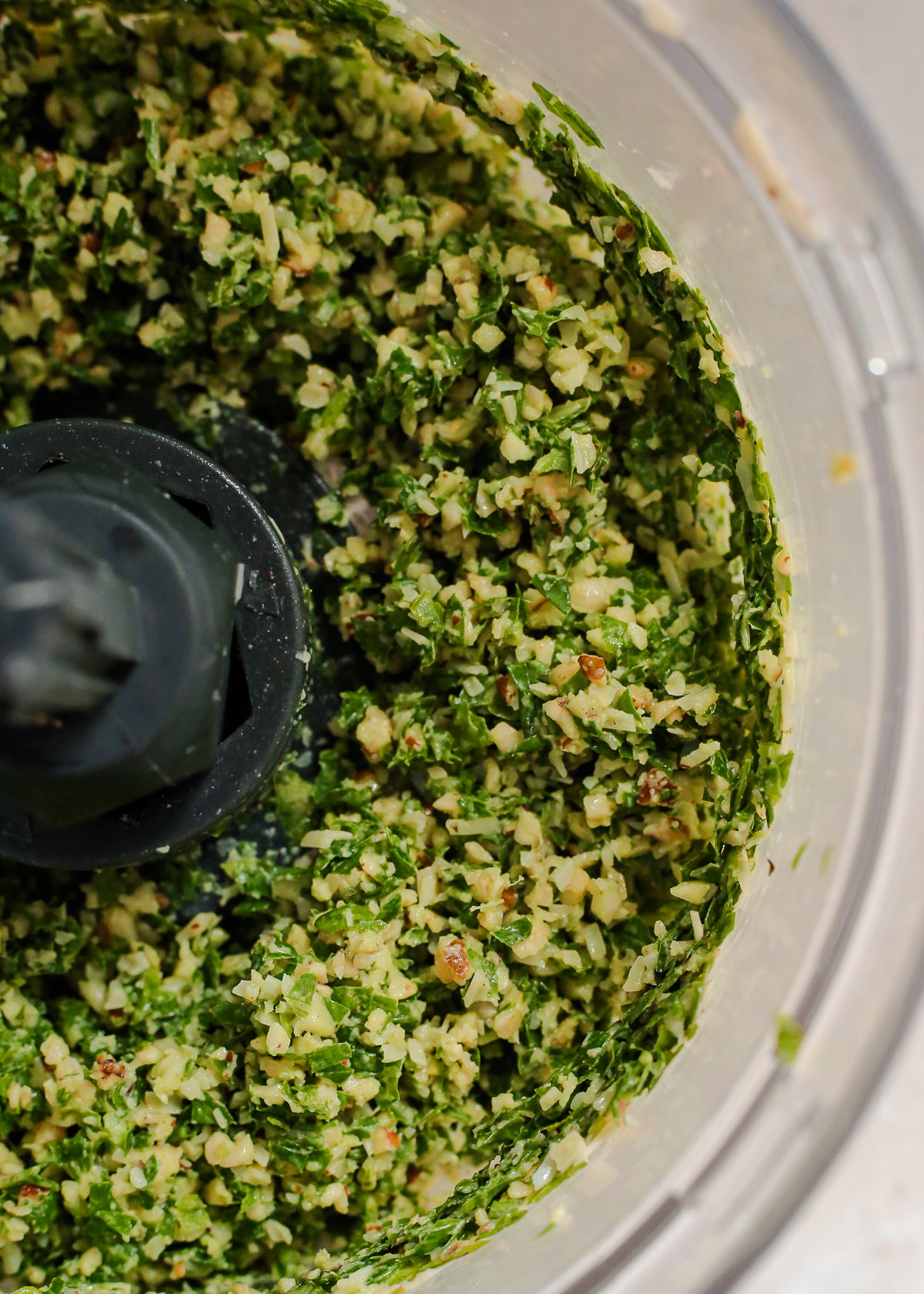 A view into the bowl of a food processor, showing a rough, chunky texture of blended basil, parmesan cheese, pine nuts, and garlic. 