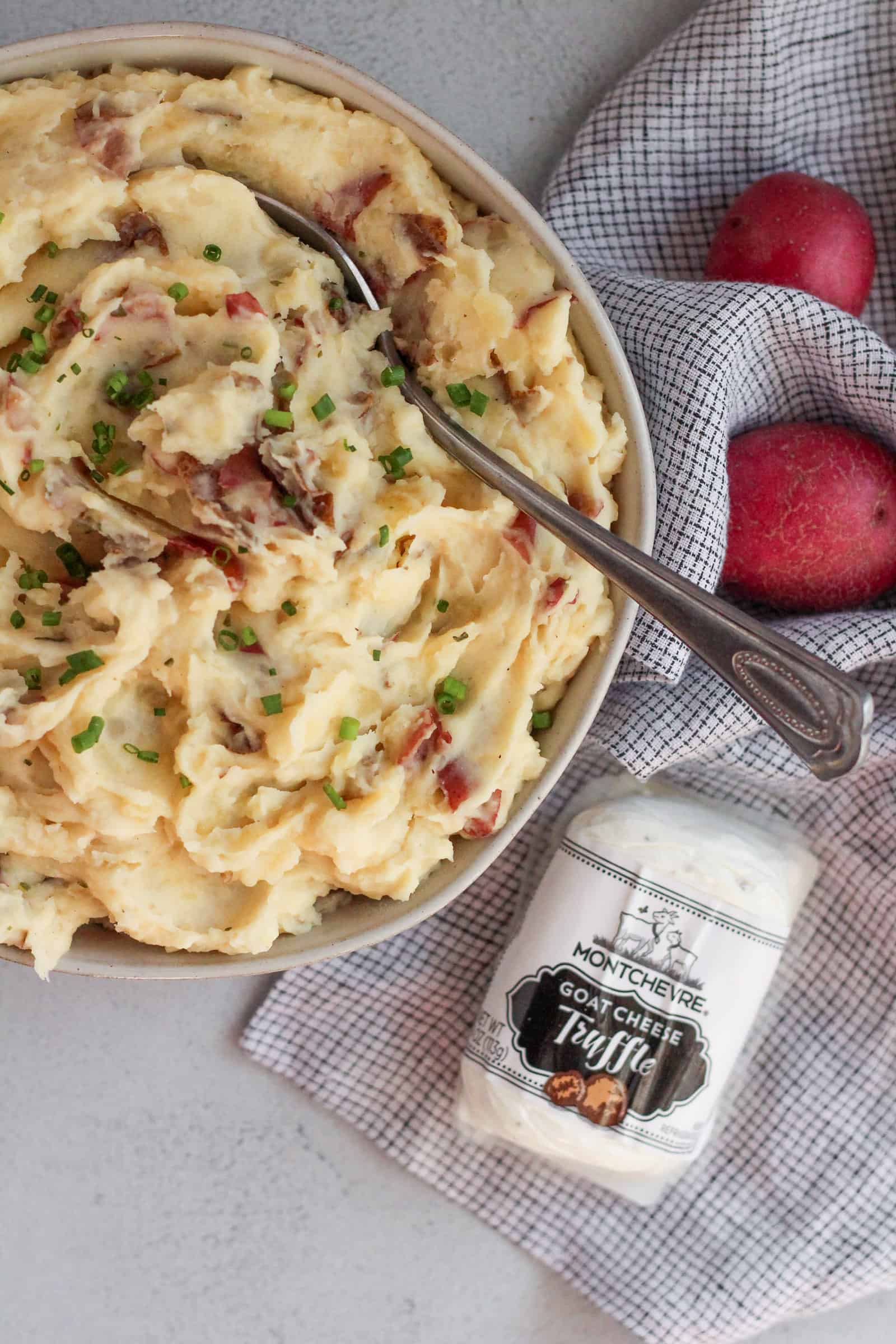 A serving bowl of truffle goat cheese mashed potatoes with a package of goat cheese and small, red skinned potatoes on a kitchen counter