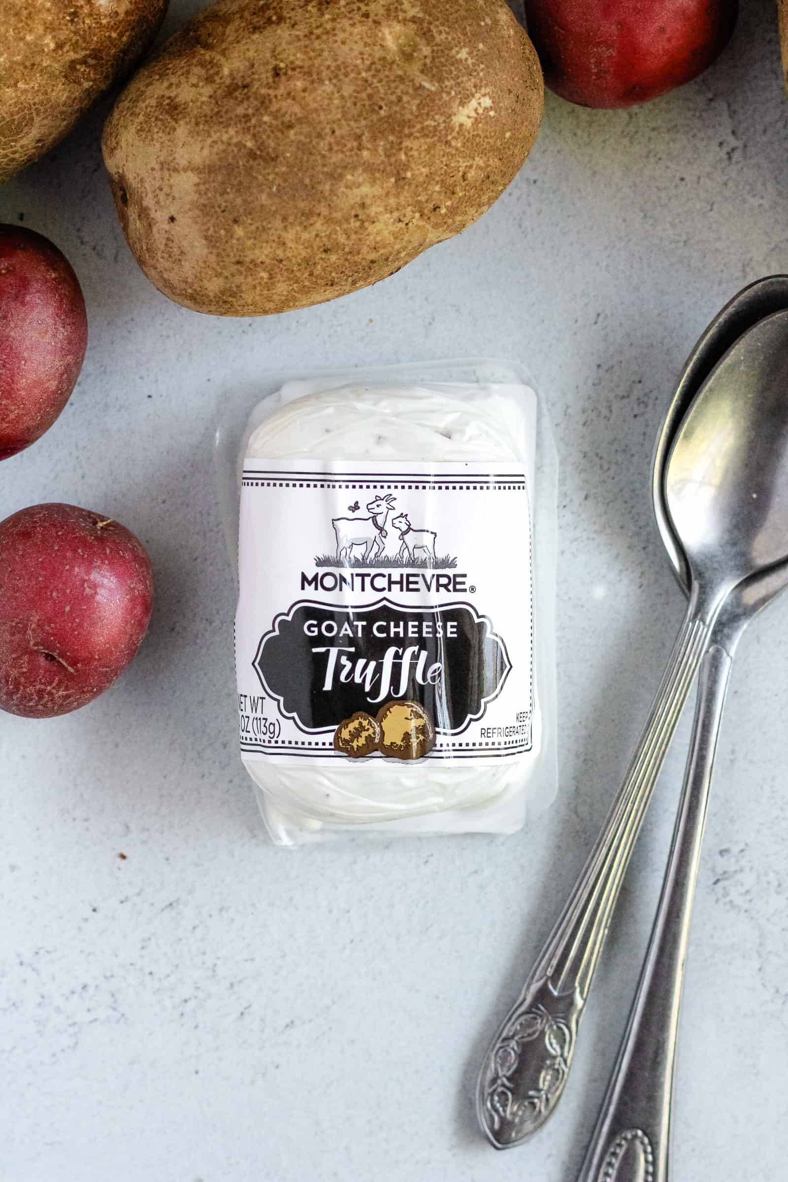 Package of creamy goat cheese on a kitchen counter, surrounded with red and Yukon potatoes and silver serving spoons