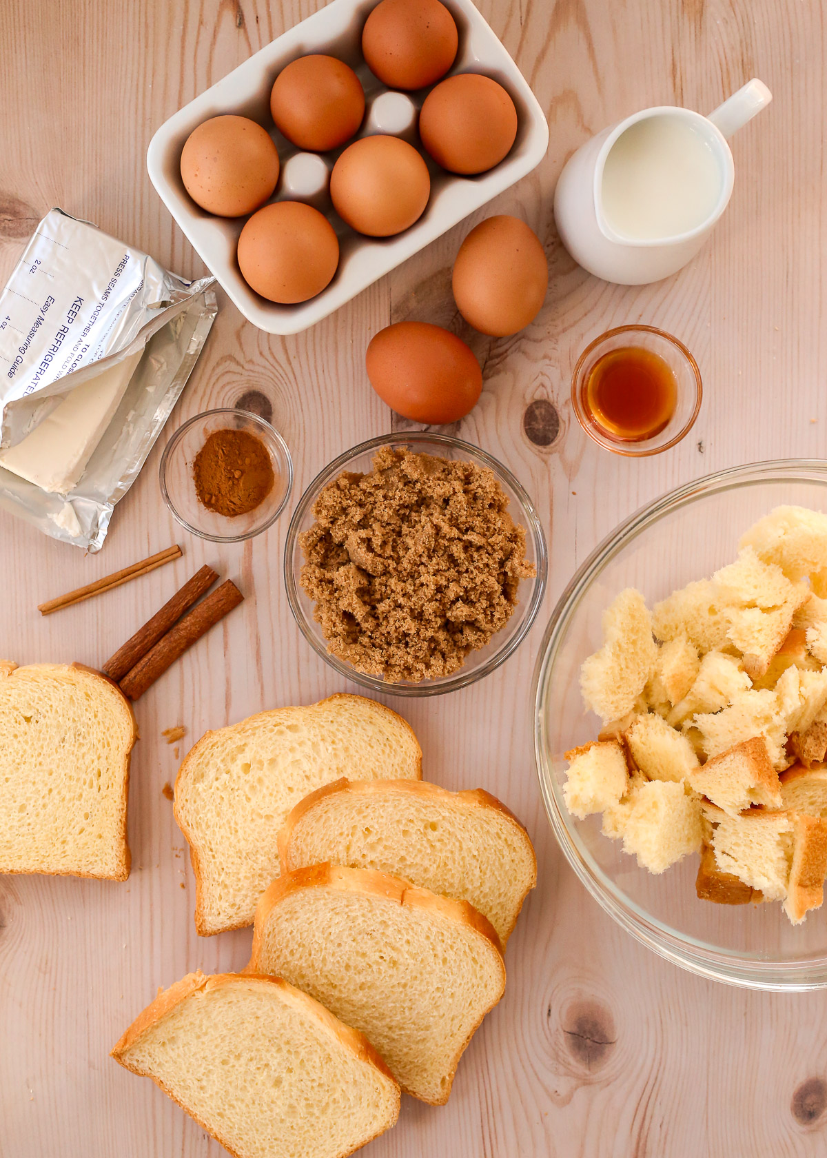An assortment of ingredients to make a baked french toast casserole, including sliced and torn brioche bread, eggs, milk, cream cheese, brown sugar, cinnamon, and vanilla.