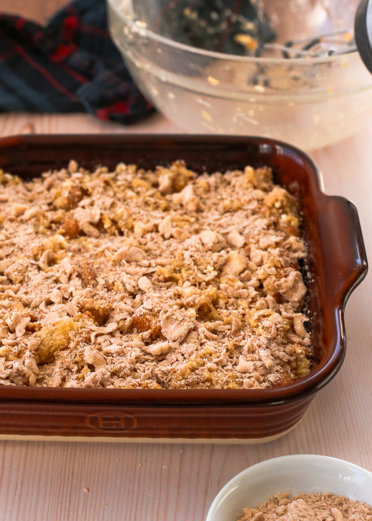 An uncooked french toast casserole, shown in a baking dish with the cinnamon streusel topping and an empty mixing bowl in the background.