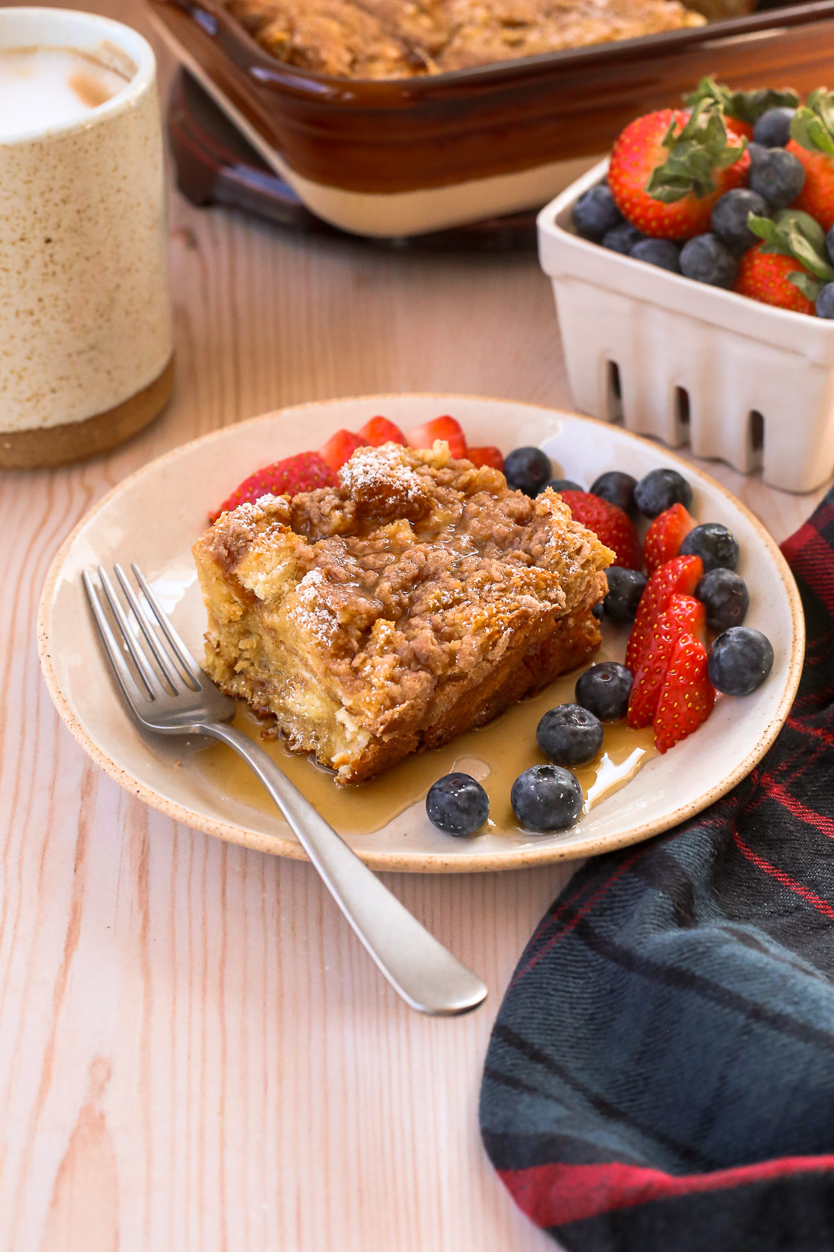 A serving of baked french toast casserole is served with sliced strawberries and fresh blueberries, topped with powdered sugar and maple syrup.