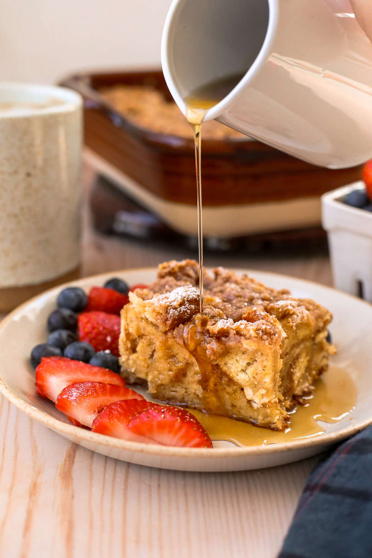 Maple syrup slowly being poured over a slice of baked french toast casserole, served on a small ceramic plate with sliced strawberries and fresh blueberries.