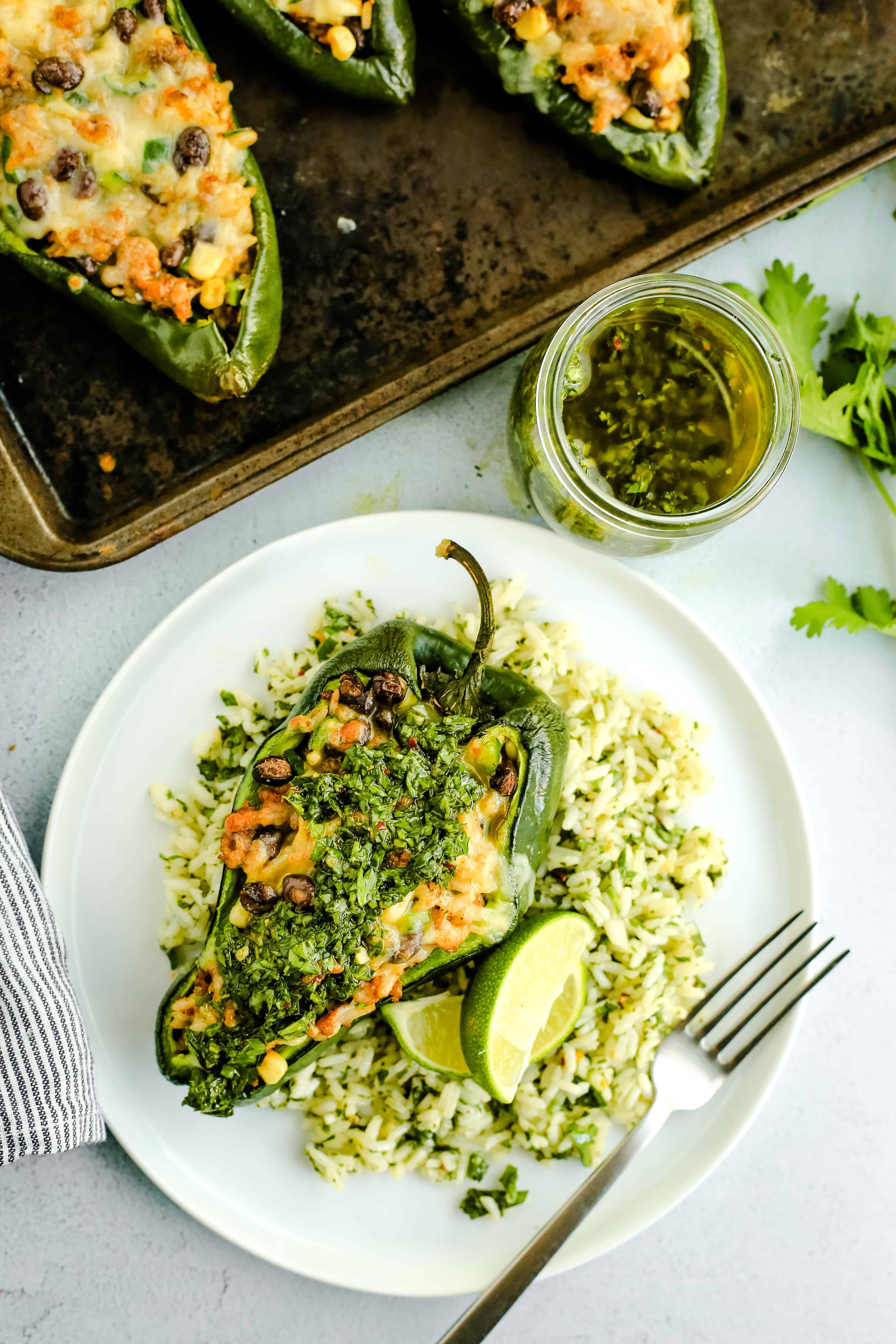 Plated and styled overhead image of Stuffed Poblano Peppers with Chimichurri Rice and Chorizo, served on a white ceramic dish with a rustic metal sheet pan and other ingredient surrounding it