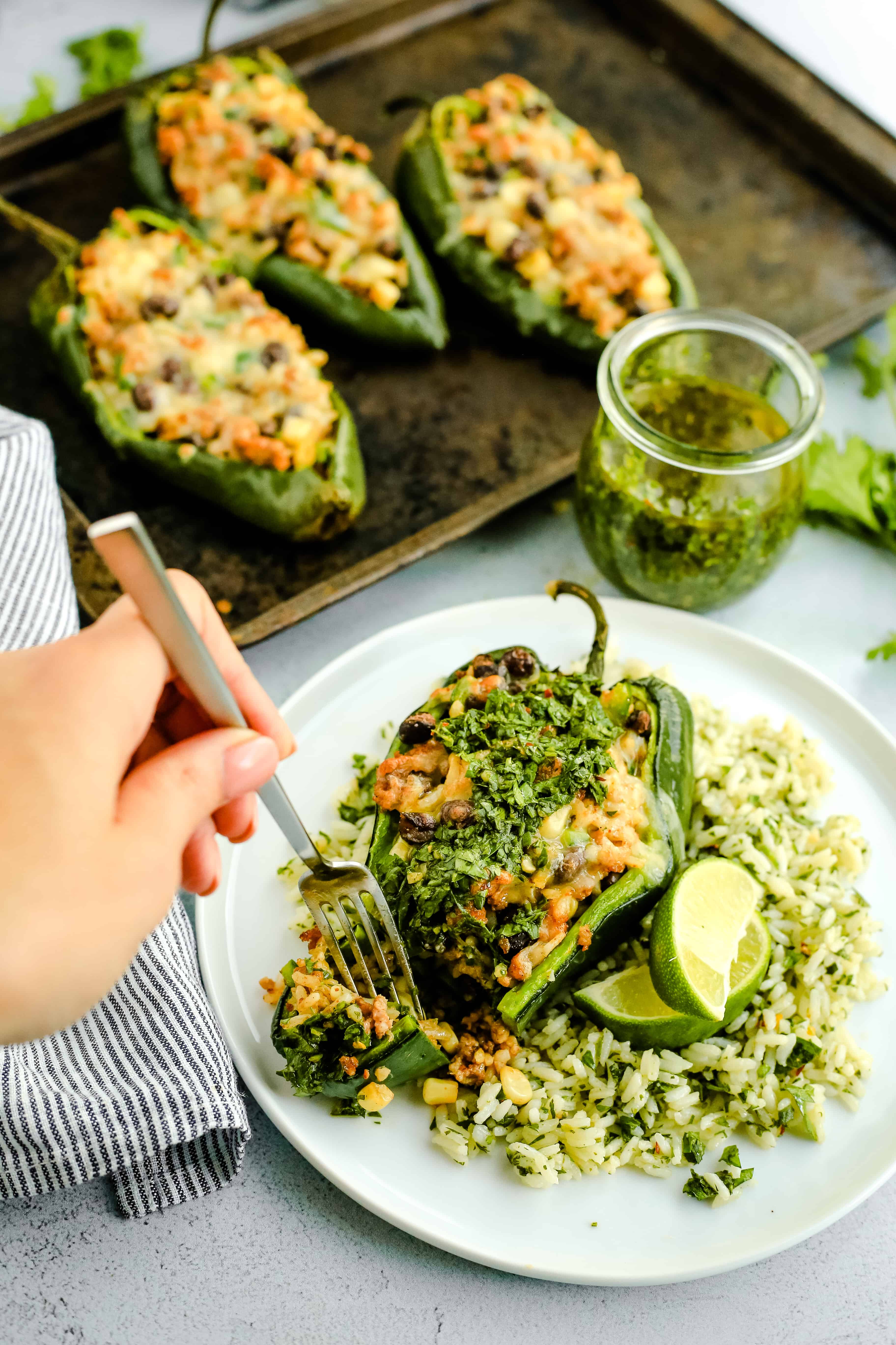 A woman's hand holds a silver fork and reached towards a plate with a stuffed poblano pepper, served on a bed of chimichurri rice with lime wedges for garnish