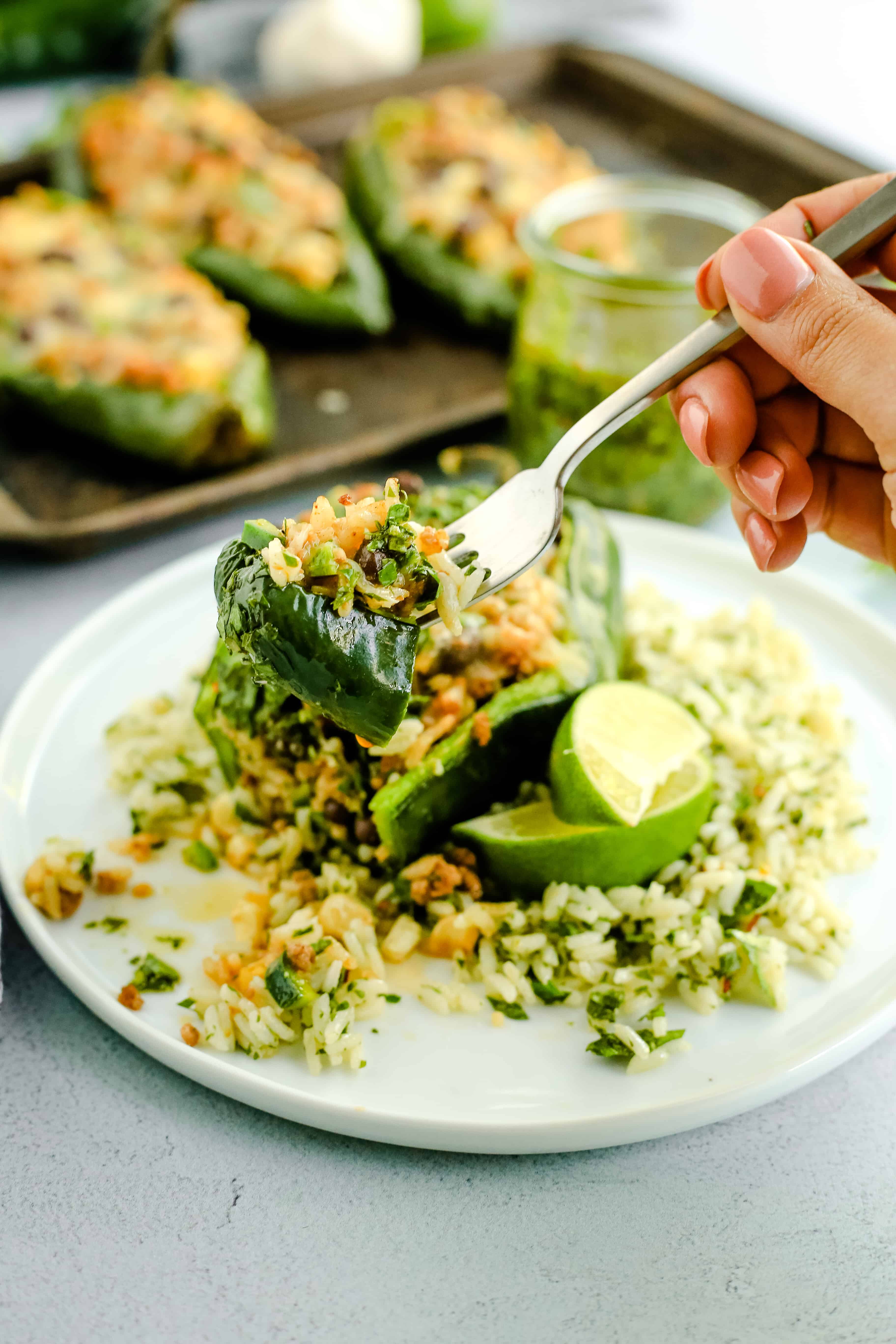 A woman's hand holds a silver fork that's balancing a full bite of stuffed poblano peppers, covered with melted cheese and chimichurri sauce while the rest of the meal is plated and within view in the background
