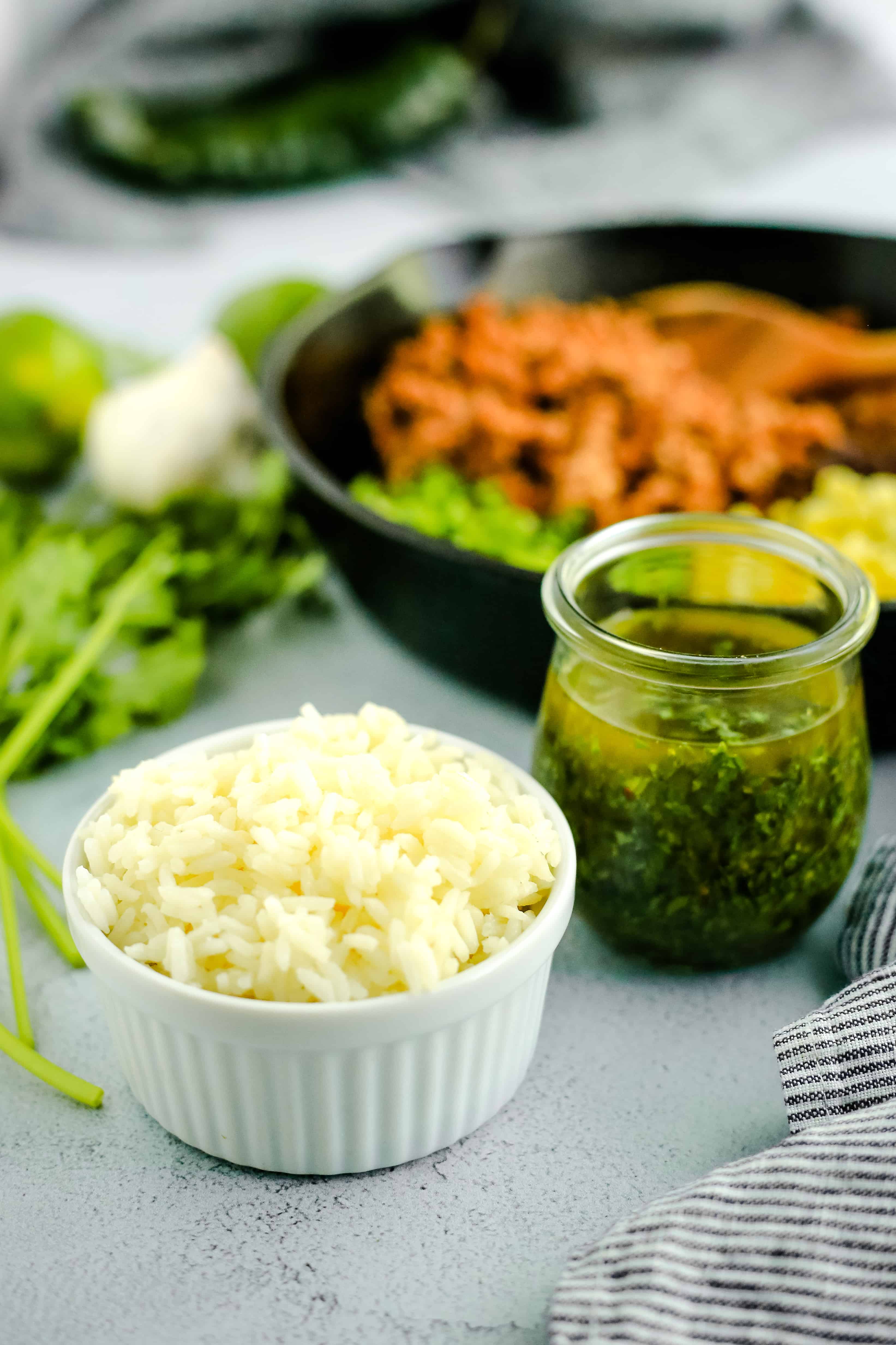 Cooked white rice in a white ramekin next to a clear glass container with freshly made chimichurri sauce, with extra herbs and greens arranged in the background on a kitchen countertop