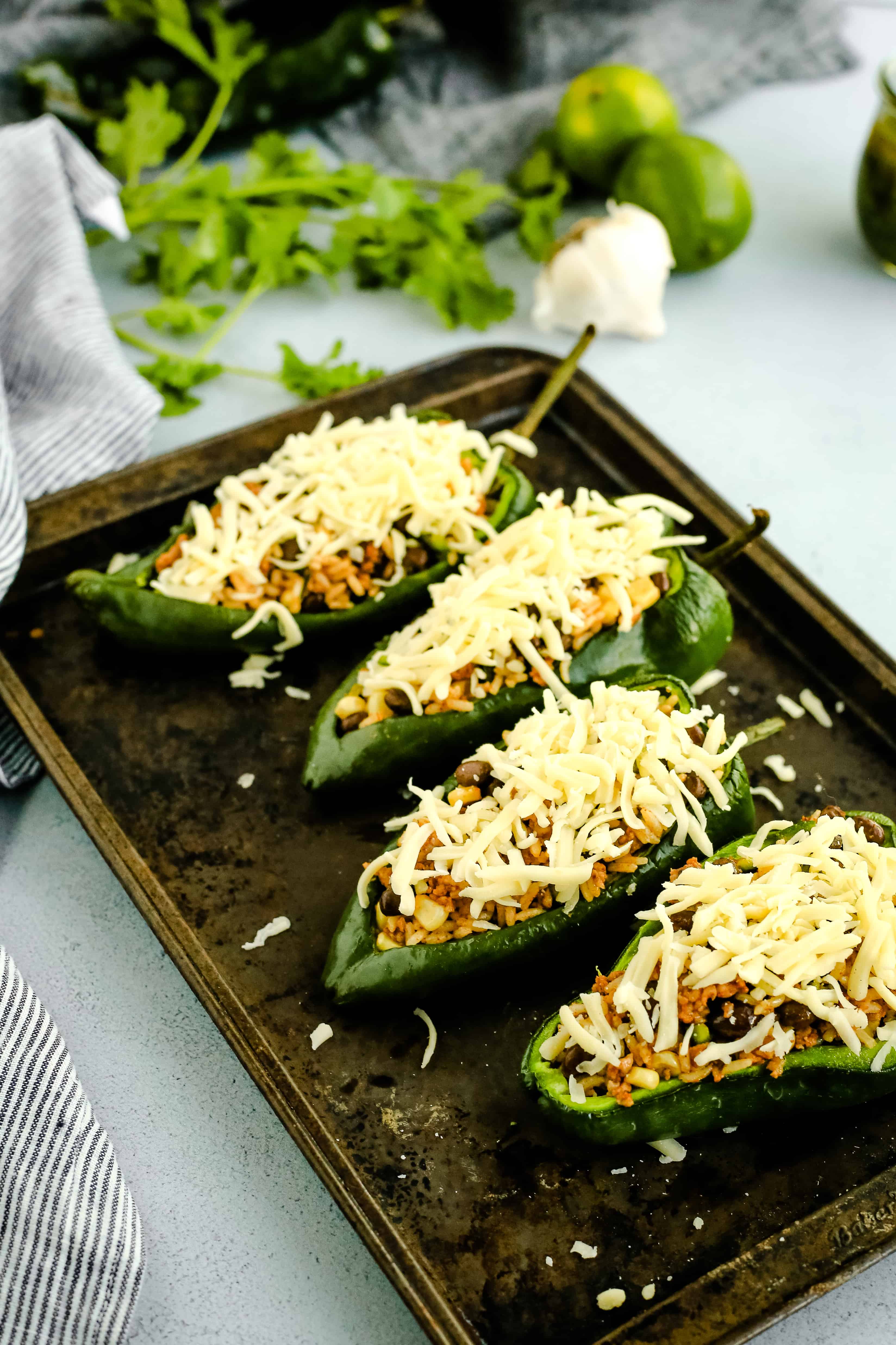Angled view of stuffed poblano peppers on a rustic metal sheet pan, topped with shredded cheese prior to being baked in the oven