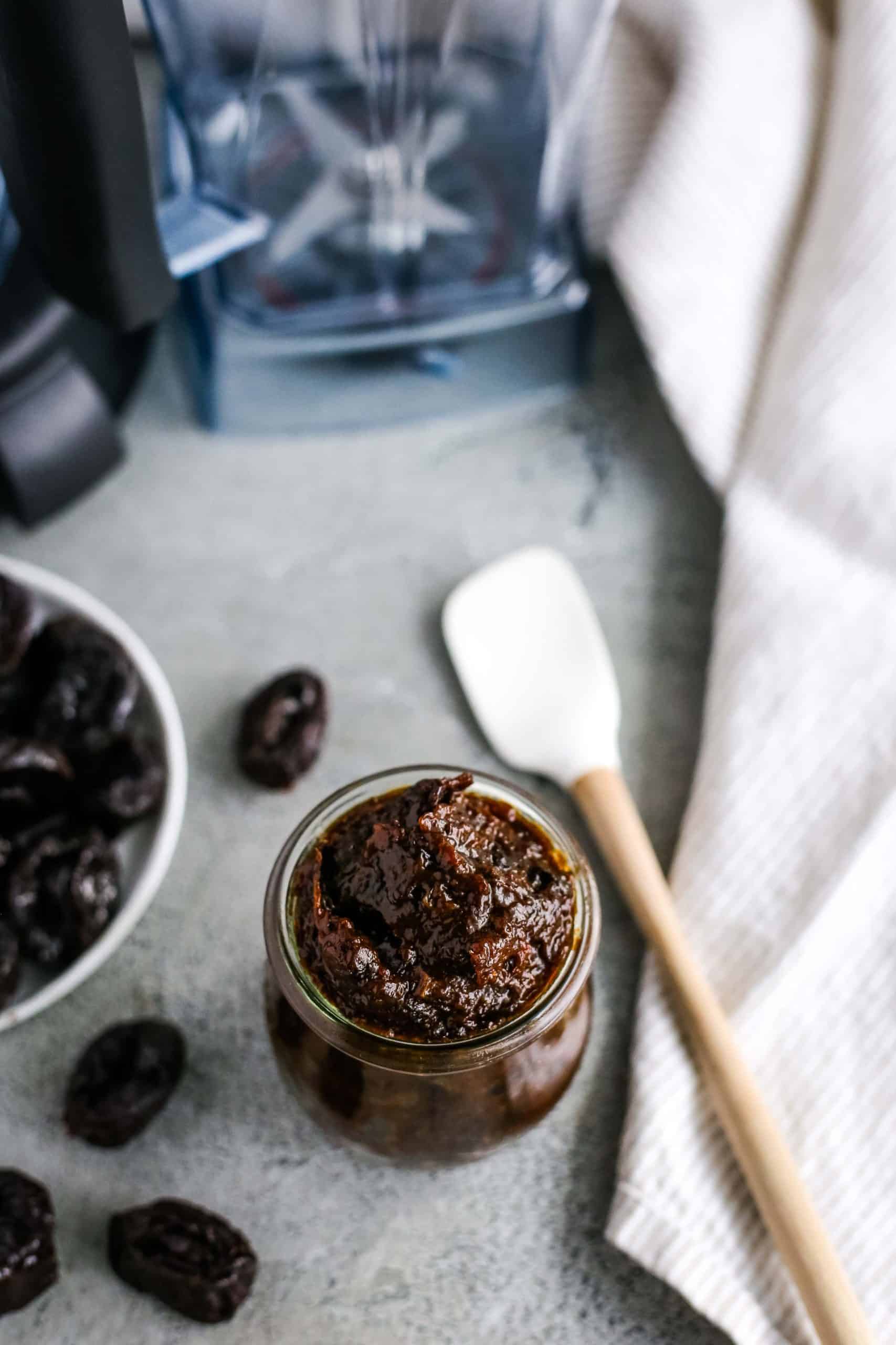 A prune puree recipe in a small glass jar with a blender, spatula, and kitchen linen placed around it on a kitchen countertop