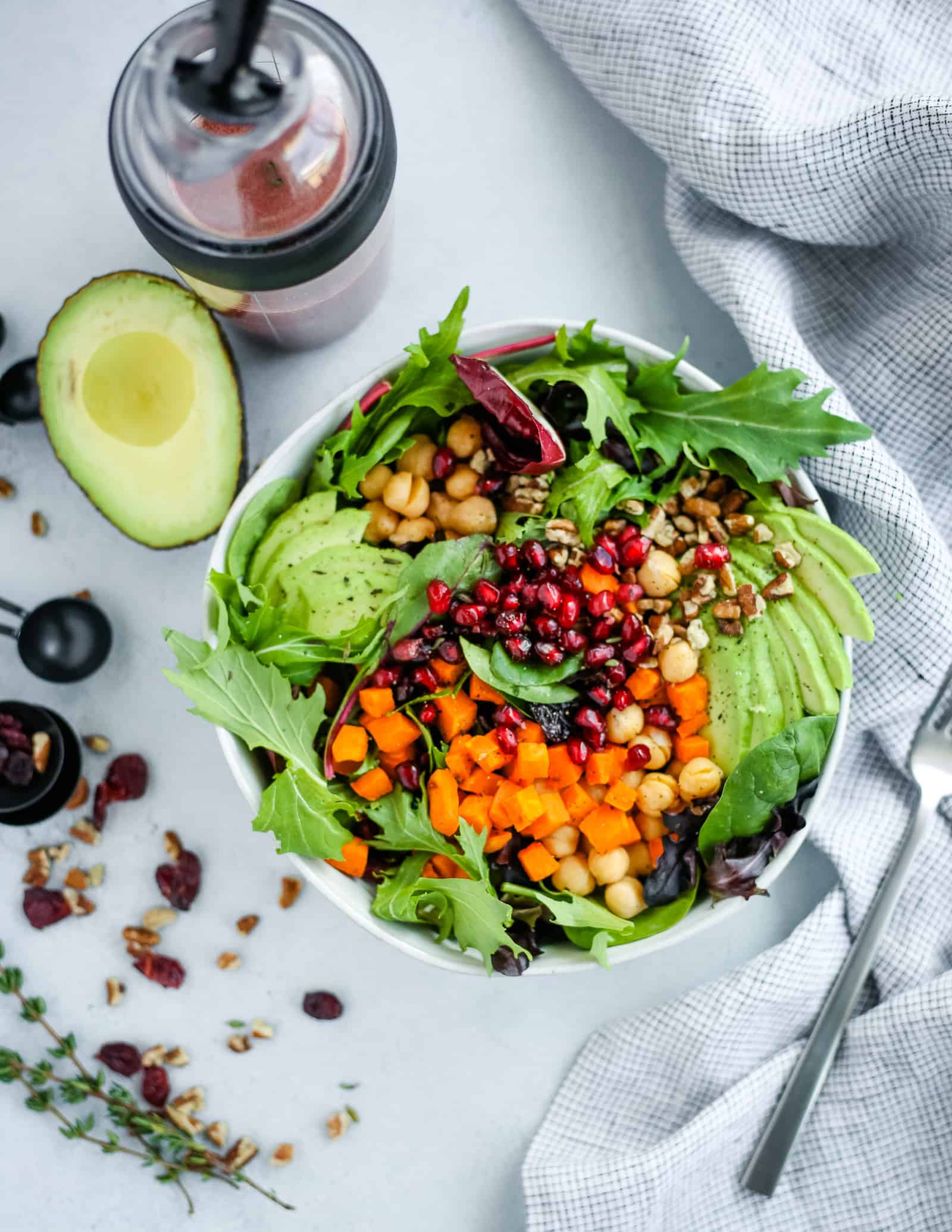 Colorful vegan pomegranate salad with fresh avocado, creamy pomegranate dressing, and a blue linen and measuring spoons, overhead view