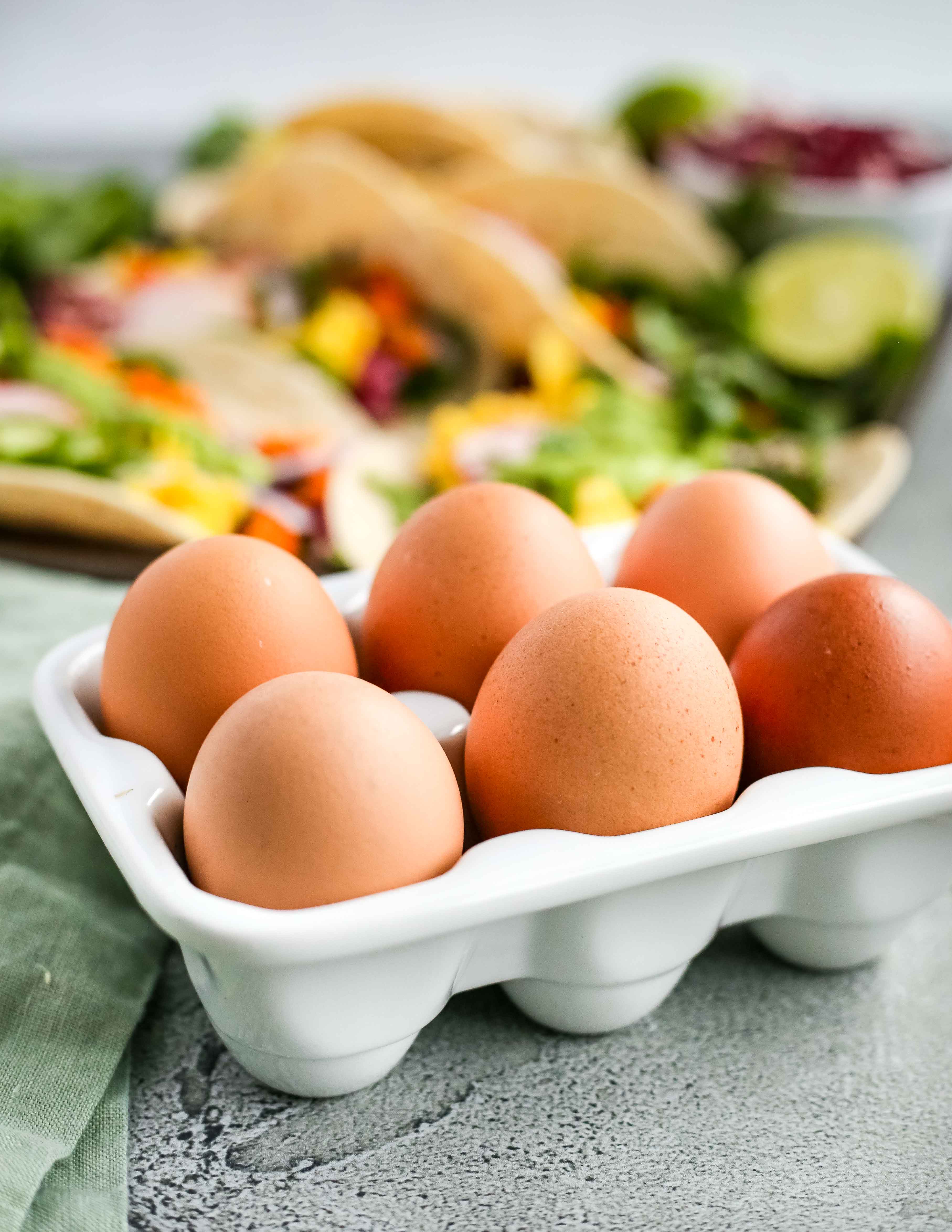 Brown eggs in a white ceramic egg holder in front of colorful breakfast tacos on a kitchen countertop