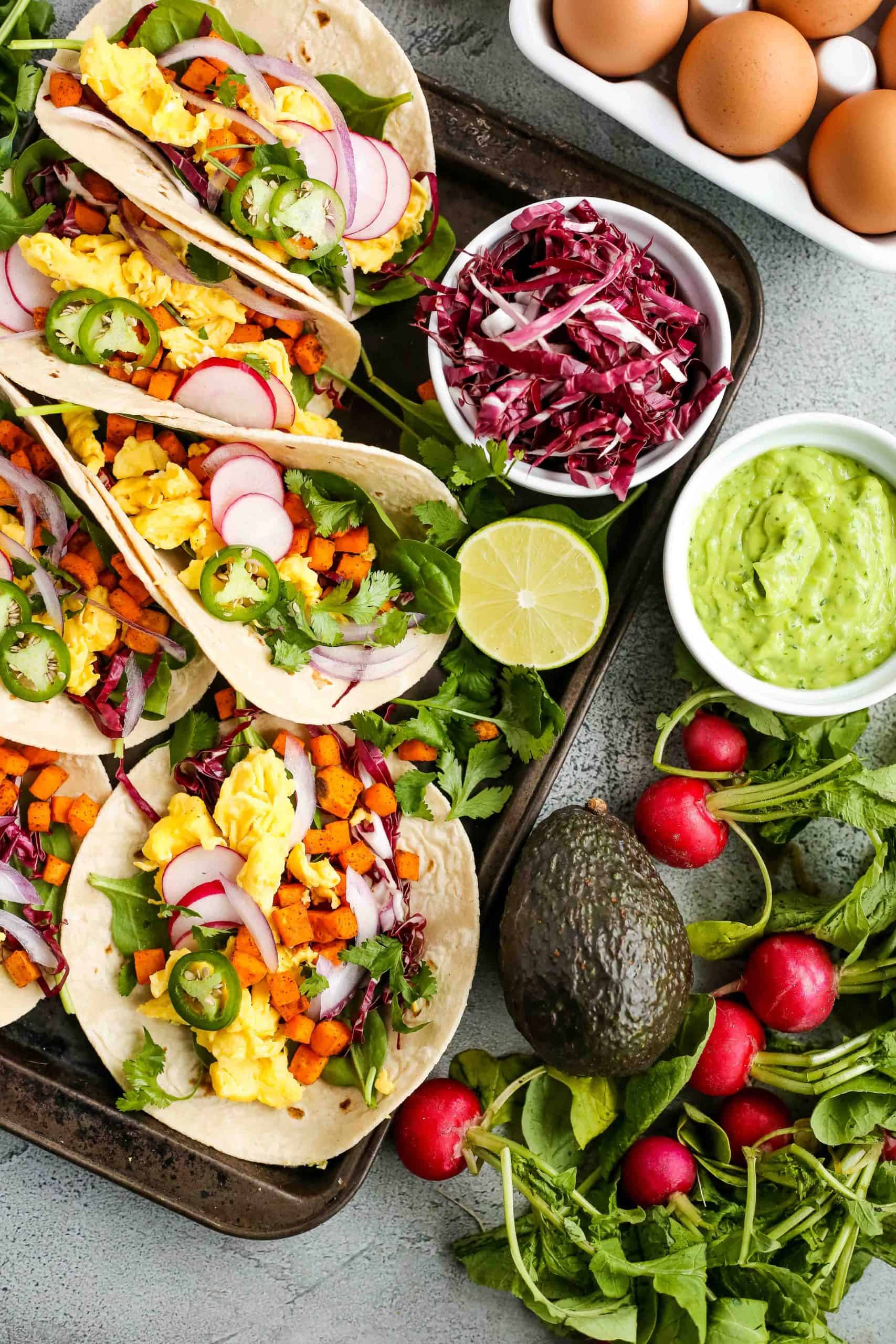 A woman's hand uses a spoon to add a creamy, thick avocado sauce onto a colorful breakfast taco filled with scrambled eggs, roasted sweet potatoes, slice jalapeños, cilantro, and red onions