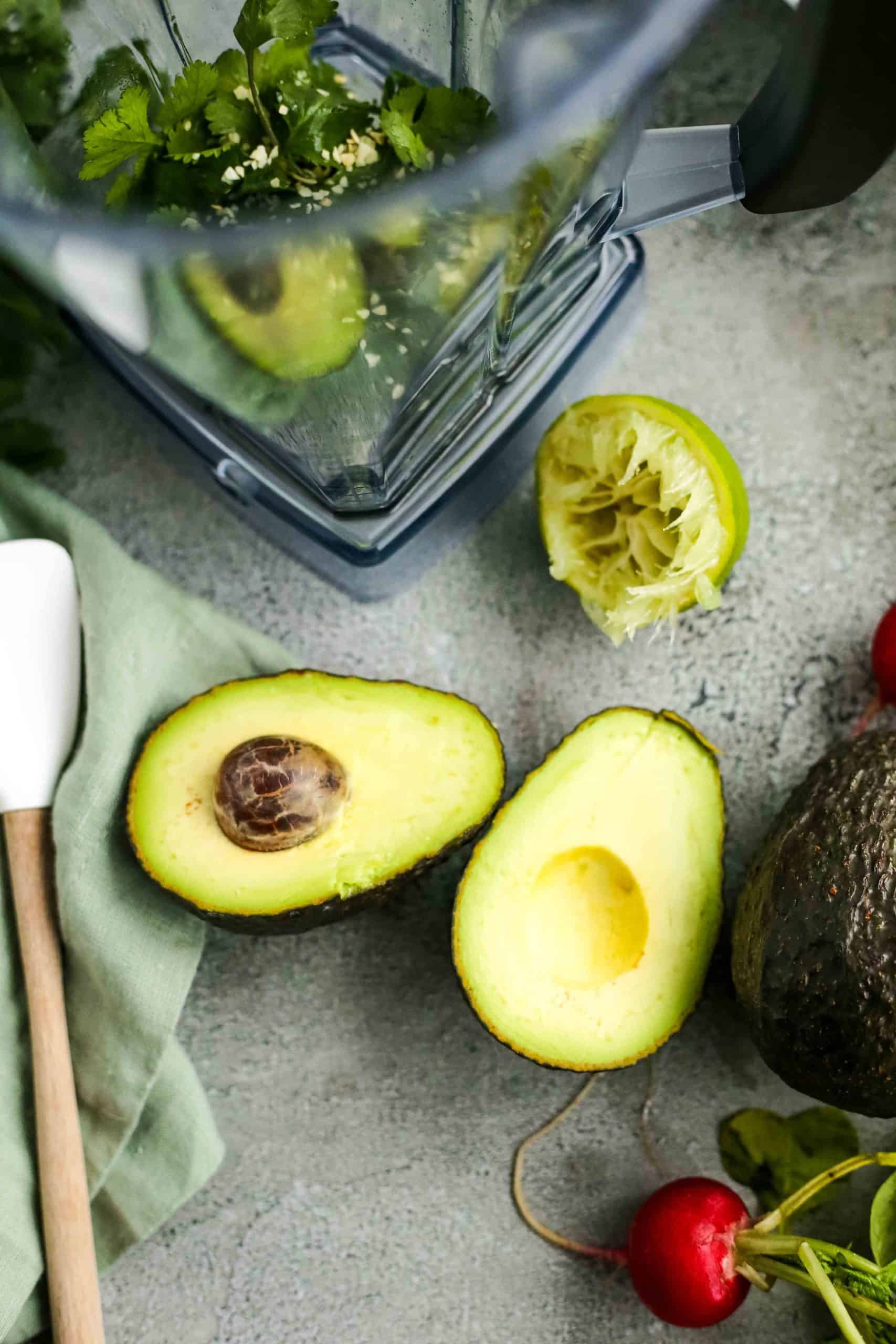 A sliced avocado lays open on a kitchen countertop near a juiced lime and blender, with cilantro and seasonings already added to the blender 