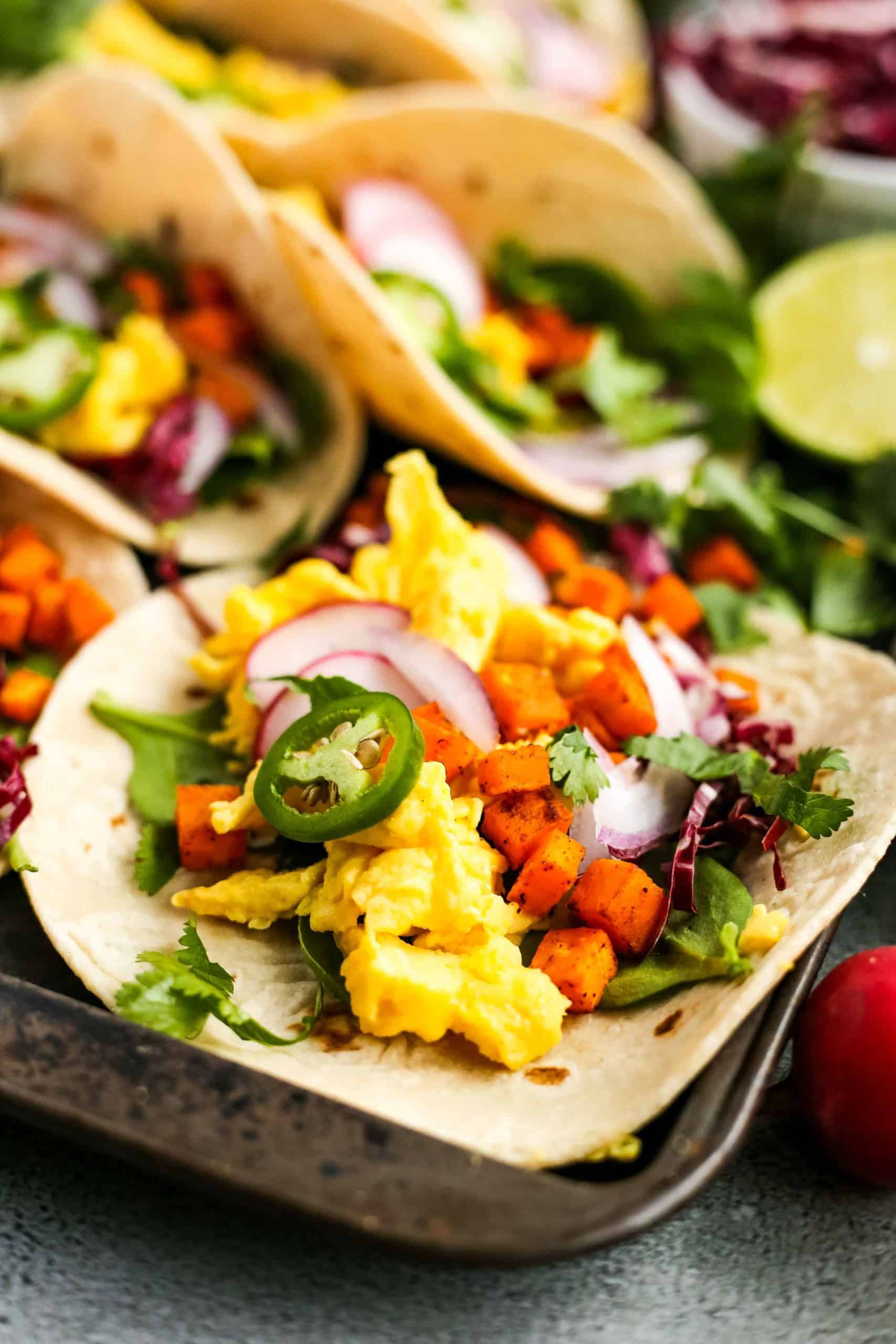 A colorful breakfast taco with scrambled eggs, sweet potatoes, and spring veggies lies open on a sheet pan, with more tacos in the background